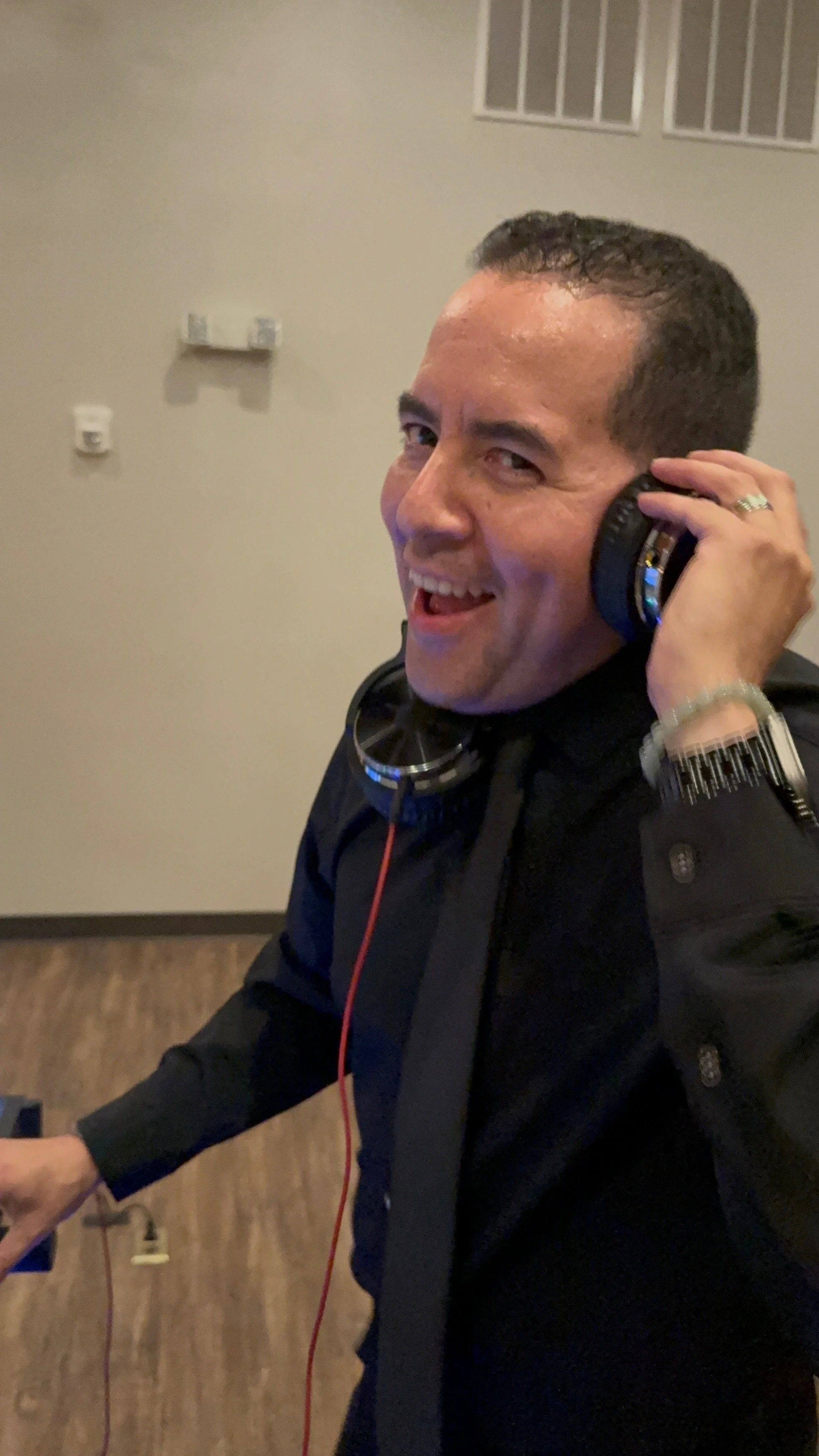 A smiling man with short dark hair wearing a black jacket and headphones around his neck, holding a device in an indoor setting.