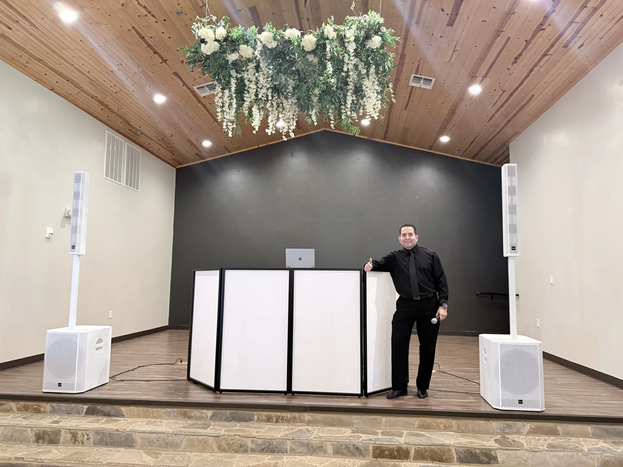 Man in black suit and tie standing on stage with white and black folding screen, large floral chandelier hanging from wooden ceiling, two white speakers on the floor, and a black chalkboard in the background.