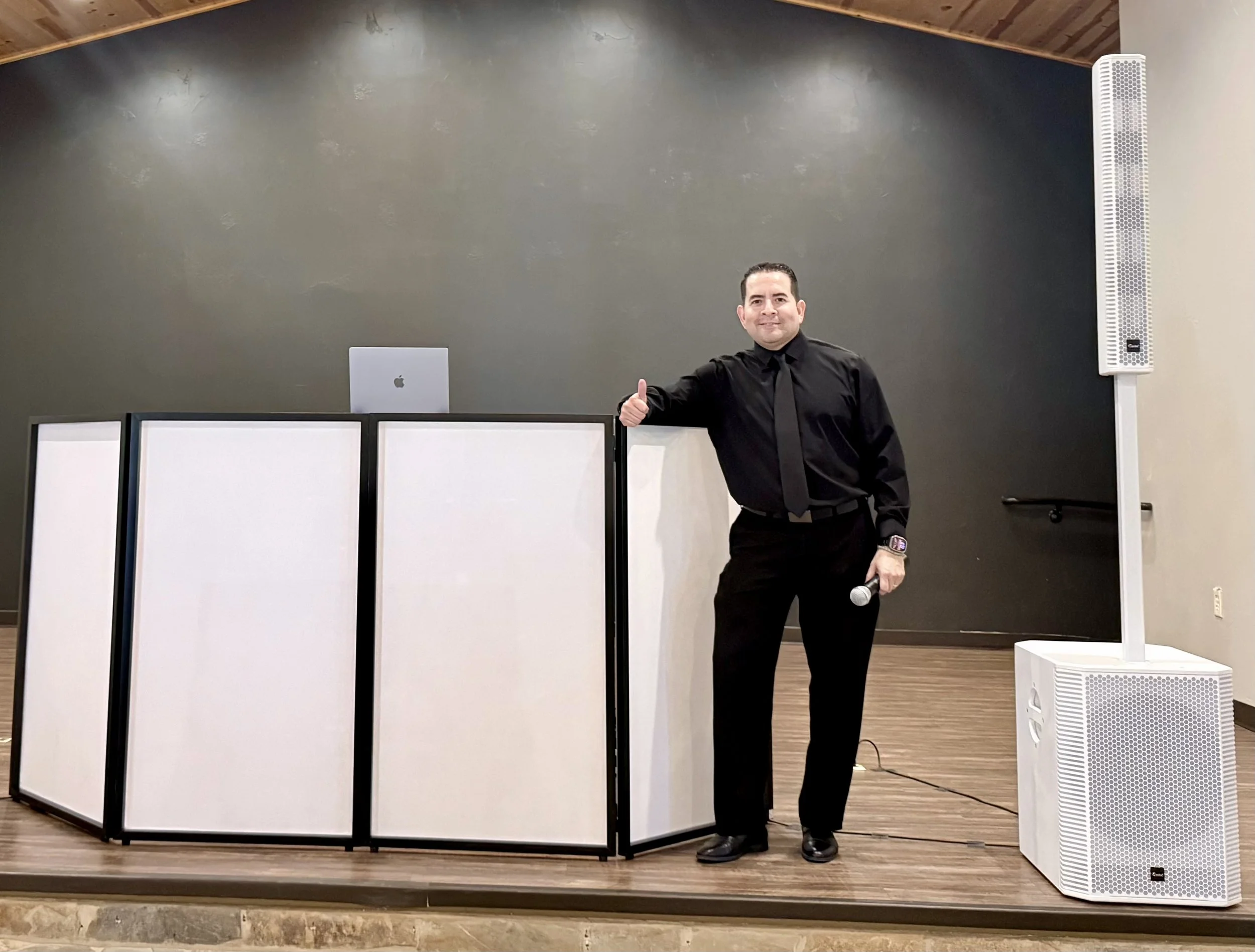 A man dressed in black with a shirt, tie, and shoes stands on a stage, giving a thumbs-up gesture. Behind him is a beige partition with a computer monitor on top, and to his right, a large white speaker or air purifier. The stage has a wooden floor, and the back wall is painted gray.