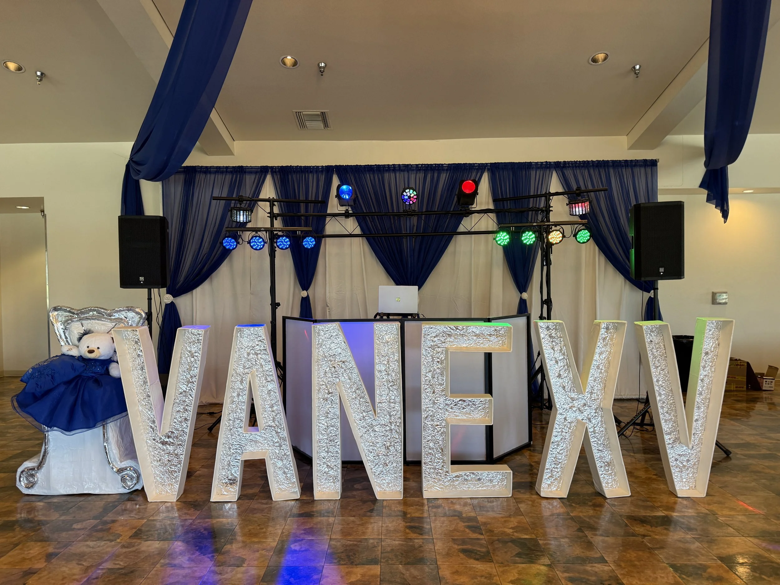 Decorated event space with large illuminated 'W' letters, small teddy bear wearing a blue dress on a chair, and a DJ booth with colorful lights, blue curtains, and speaker systems.