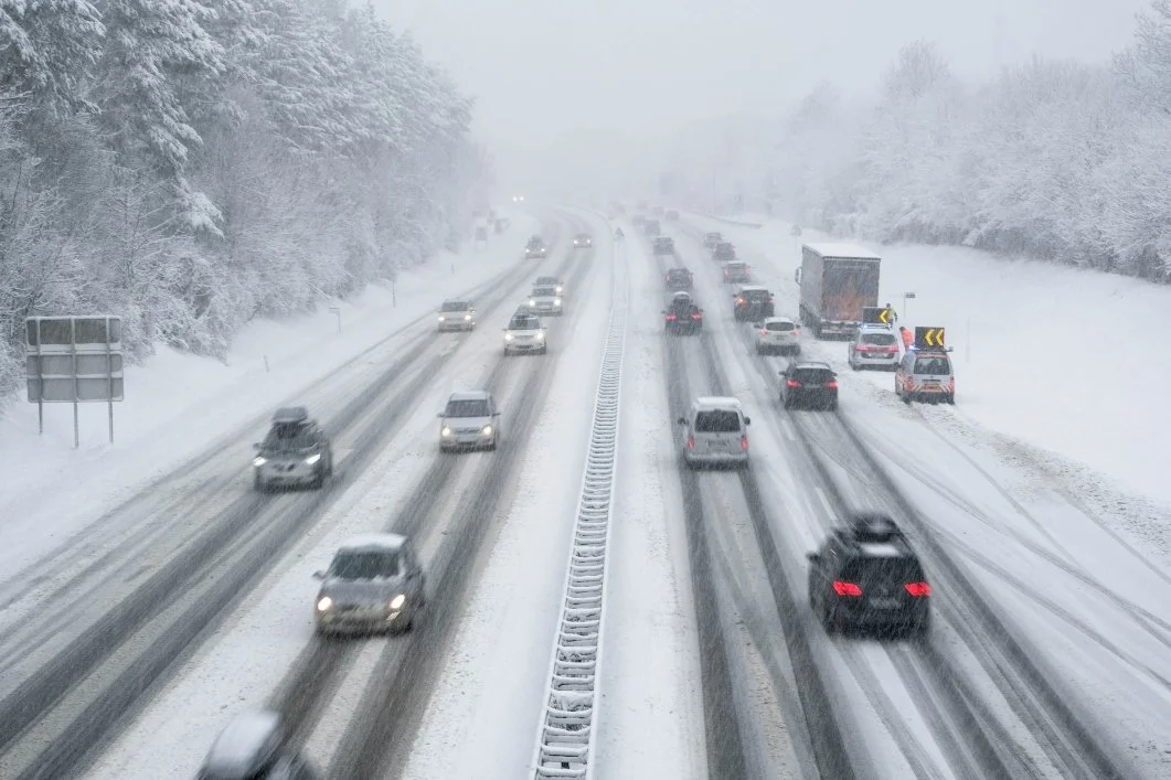 Snow-covered highway with moving and stationary cars, and snow plow clearing the road, surrounded by snow-covered trees.