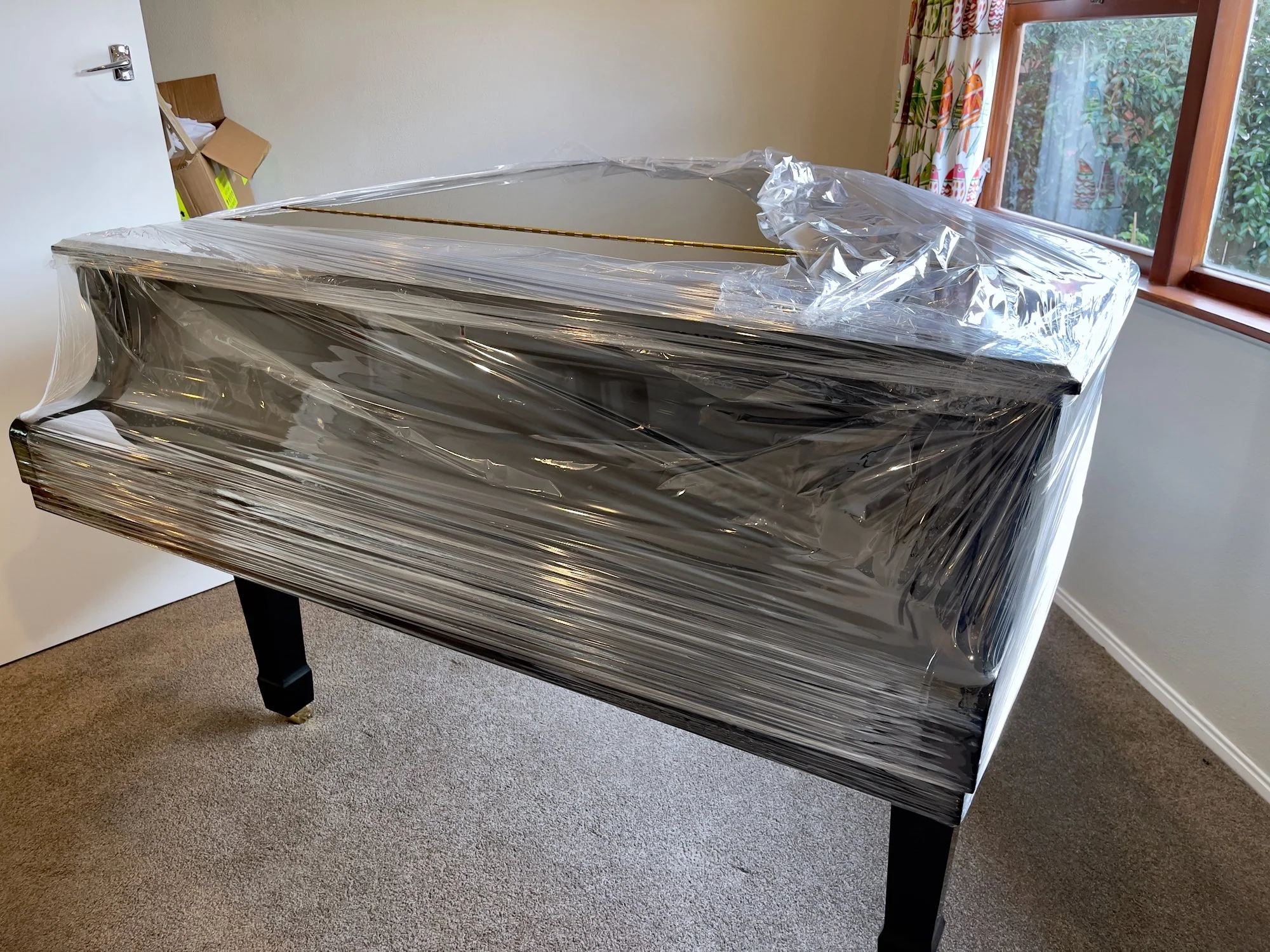 Grand piano wrapped in plastic film, situated on a room carpeted floor near a window with colorful curtains.