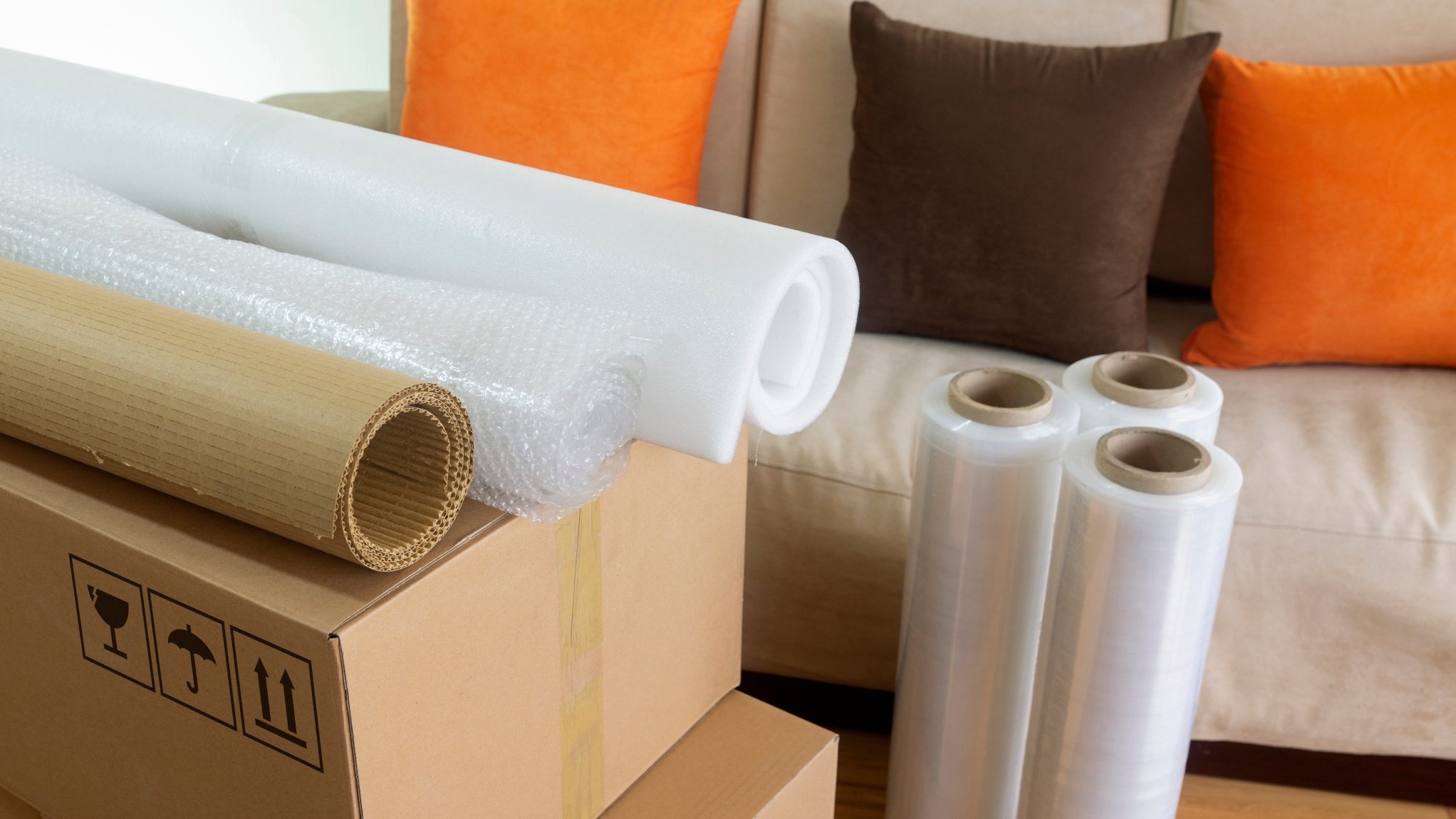 Rolls of bubble wrap, packing paper, and plastic stretch wrap on a cardboard box with moving symbols, and a beige sofa with orange and brown pillows in the background.