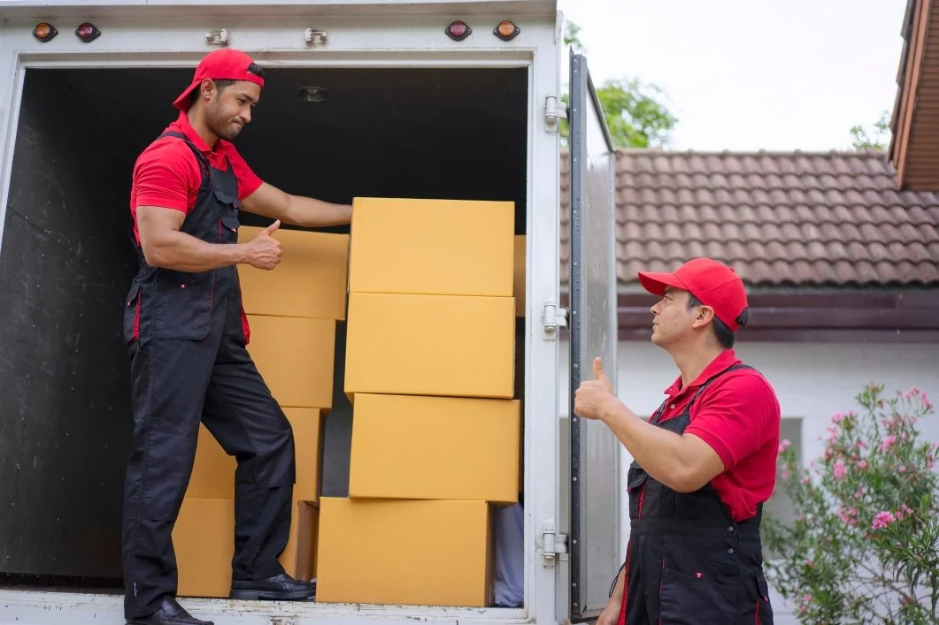 Two delivery workers, wearing red caps and red shirts with black overalls, unload yellow boxes from a delivery truck. One is inside the truck, giving a thumbs-up, while the other is outside, giving a thumbs-up back.