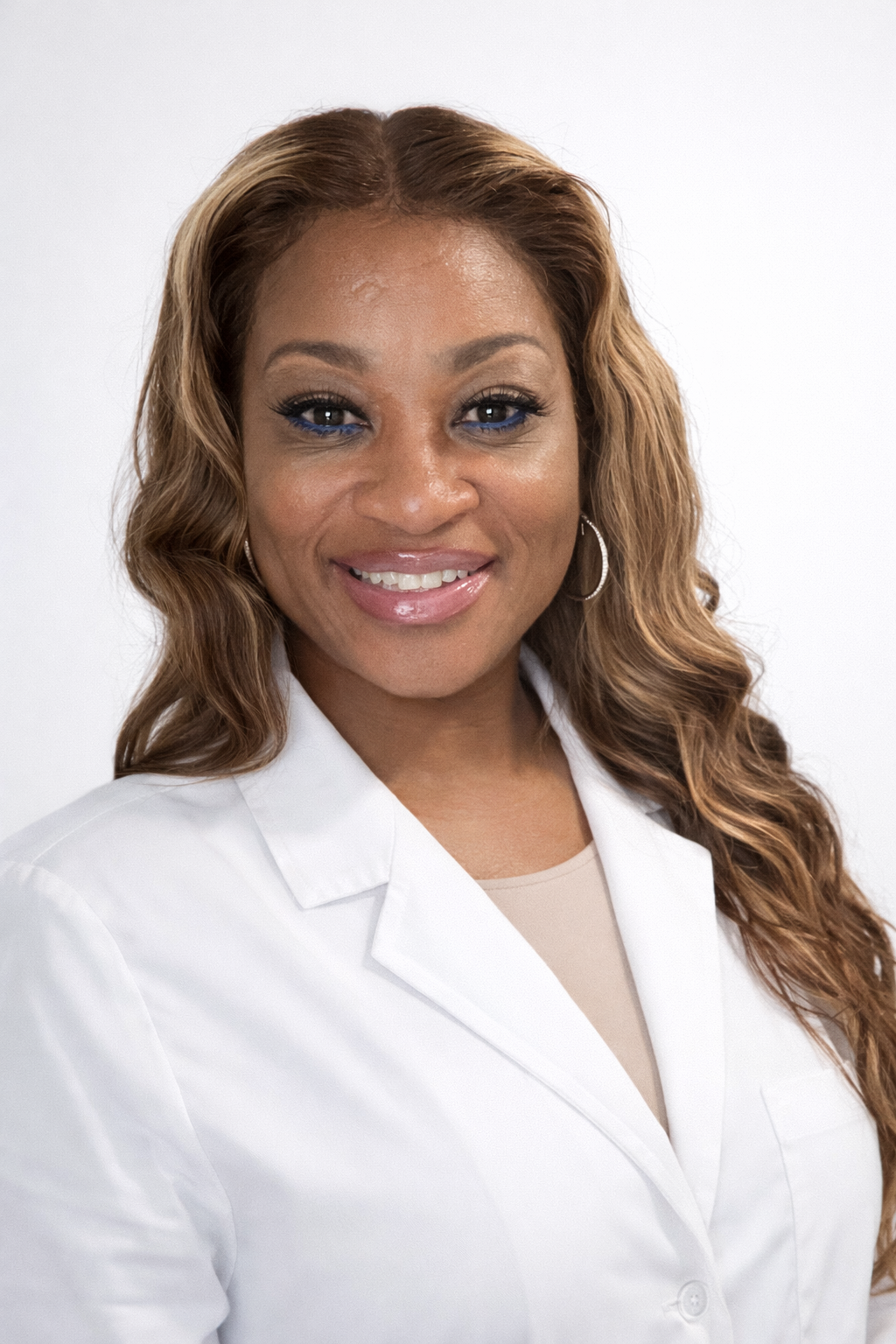 A smiling woman with long, wavy, light brown hair, wearing a white lab coat and hoop earrings, against a plain white background.