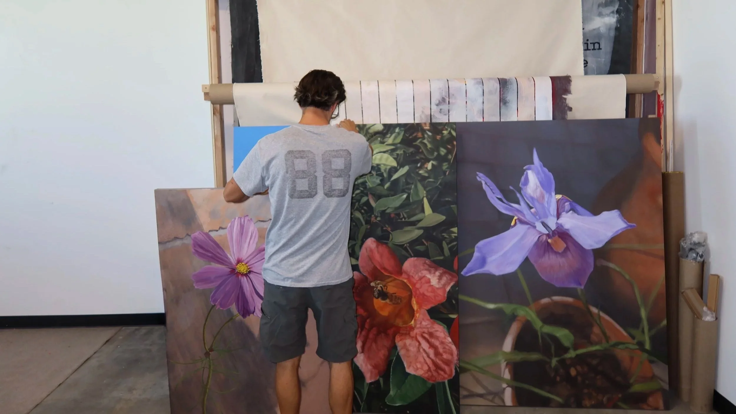 An artist arranging large floral paintings in a studio, with a person facing away from the camera, wearing a gray T-shirt and shorts.