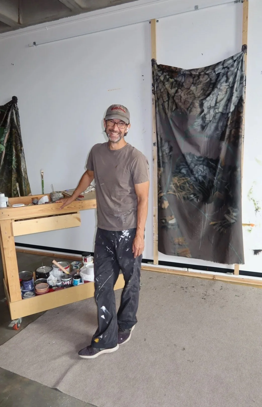 A man with glasses, a cap, and paint-splattered clothes smiling while standing in an art studio, surrounded by painting supplies. There is a large, colorful, abstract painting on the wall behind him.