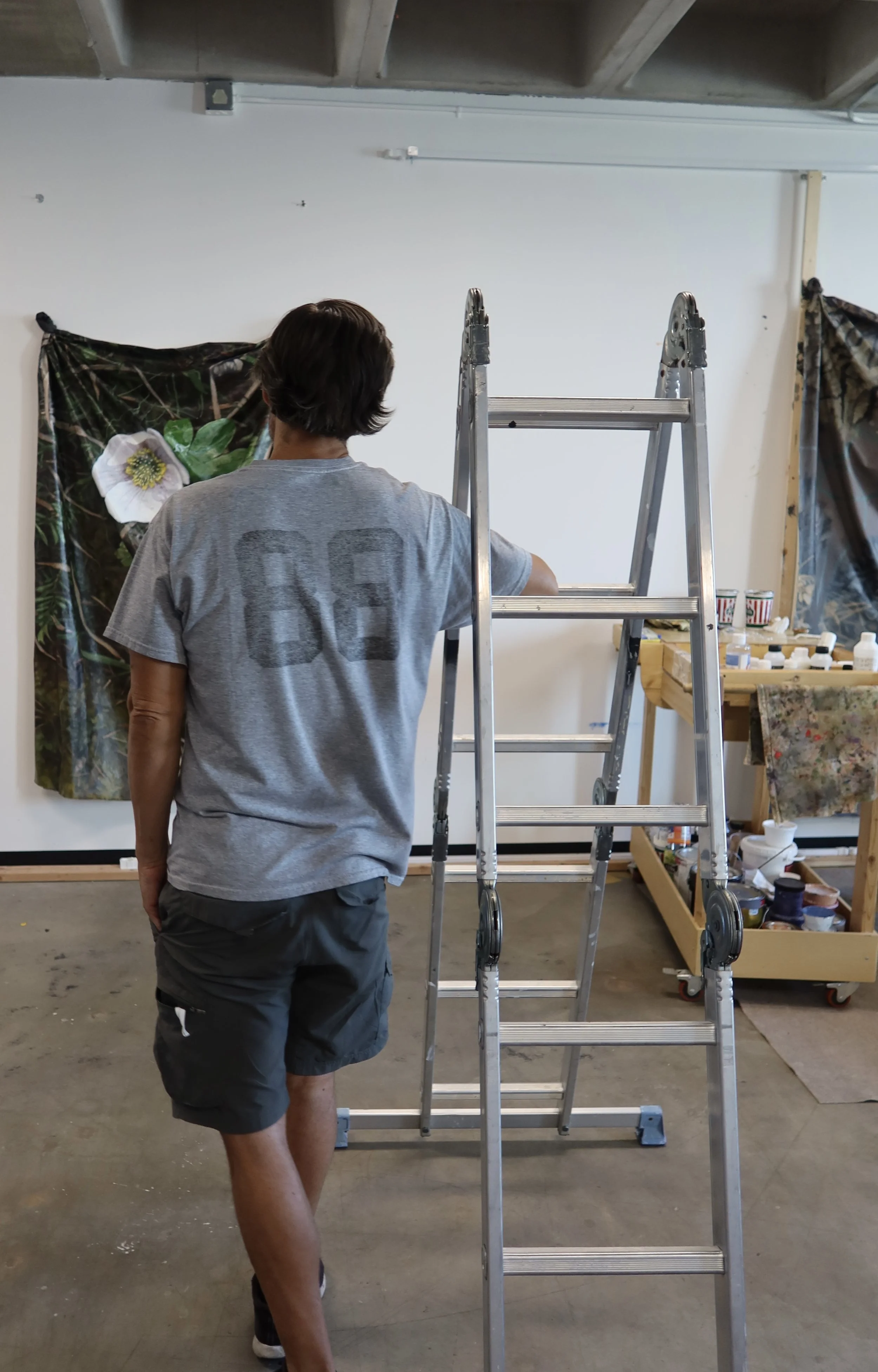 A man with dark hair wearing a gray t-shirt and dark shorts walking in an art studio. There is a ladder in the middle of the room, and a large floral painting hanging on the wall to the left. On the right, there is a wooden table with art supplies and a fabric covered partially with paint splatters.