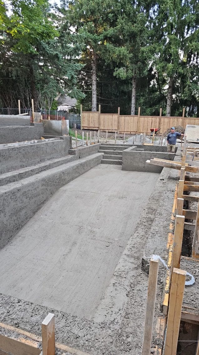 A backyard construction site with newly poured concrete for a swimming pool, surrounded by wooden framing and green trees in the background.