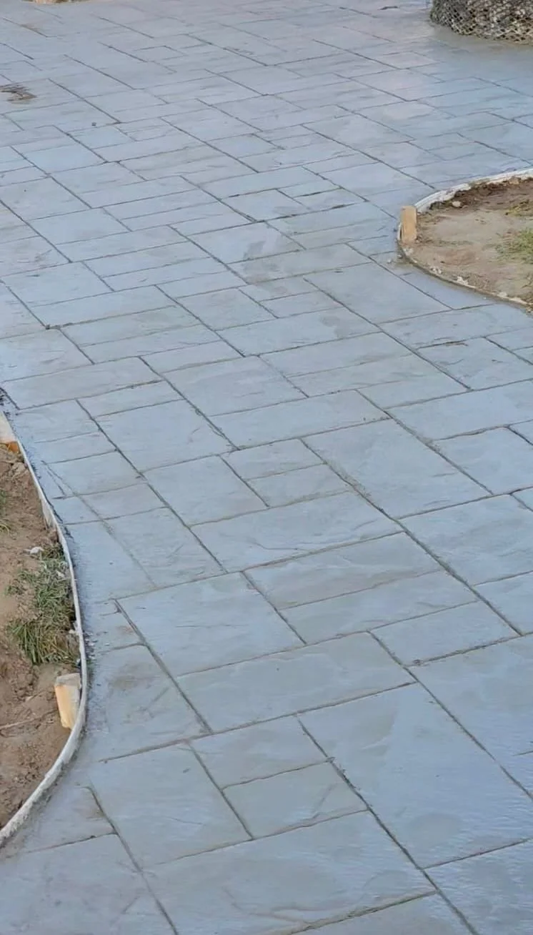 Gray stone pathway under construction, with some areas bordered by wooden stakes, adjacent to patches of dirt and grass.