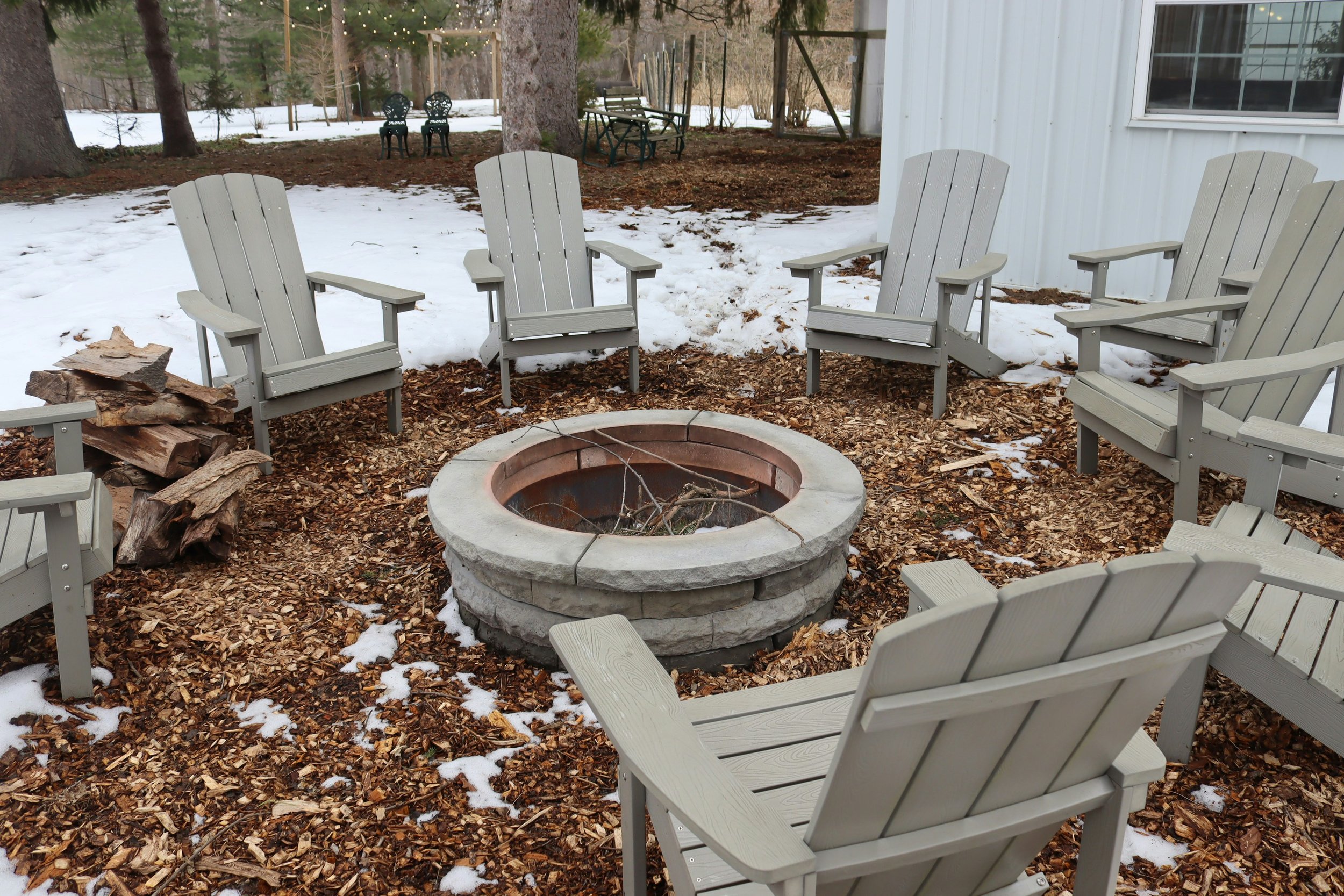 Outdoor fire pit circle surrounded by gray Adirondack chairs, with wood and snow on the ground, and trees in the background.