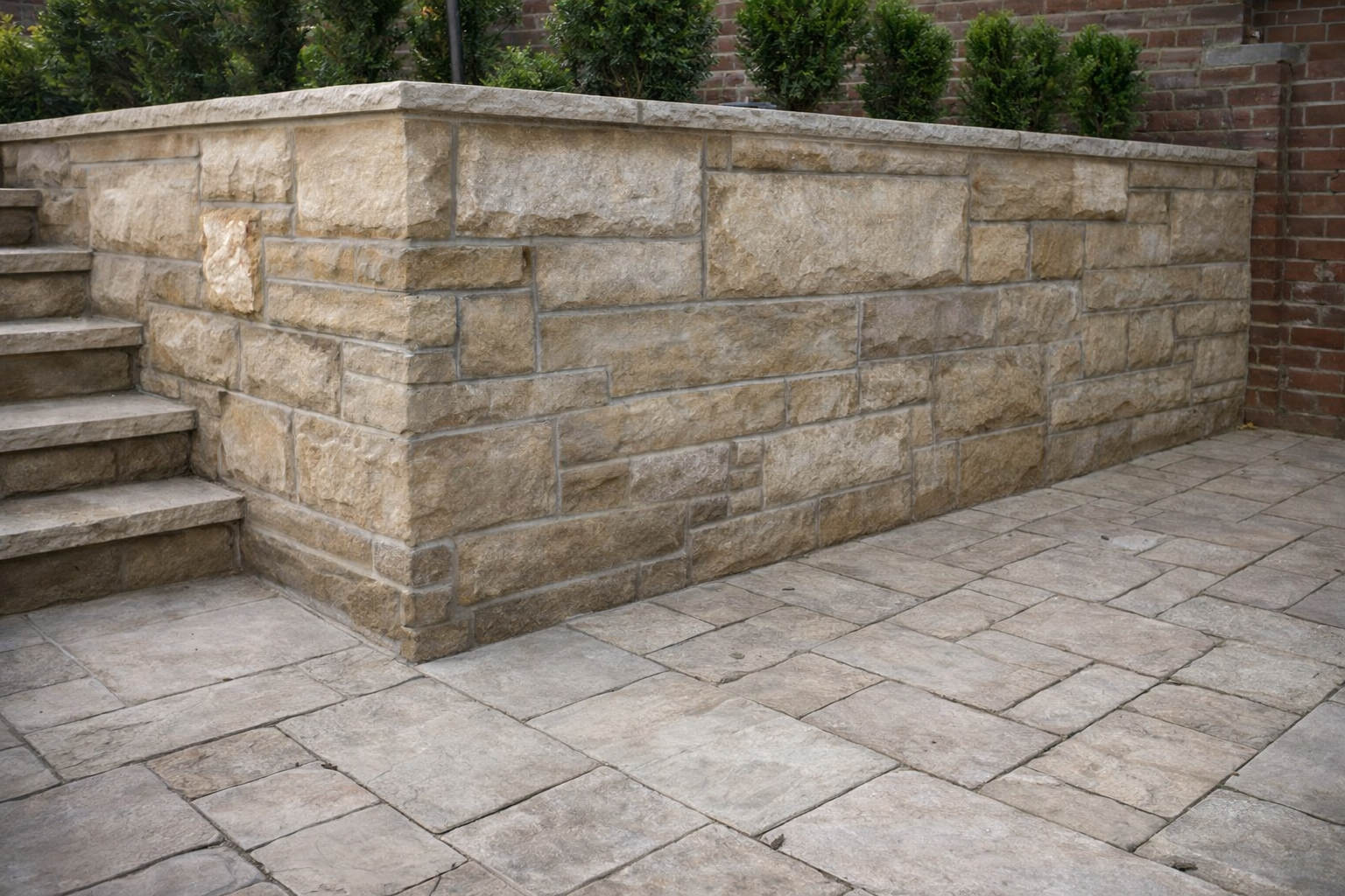 A stone retaining wall with steps on the left side, made of beige-colored stones, with a brick wall and green bushes in the background, and the ground covered with large stone tiles.