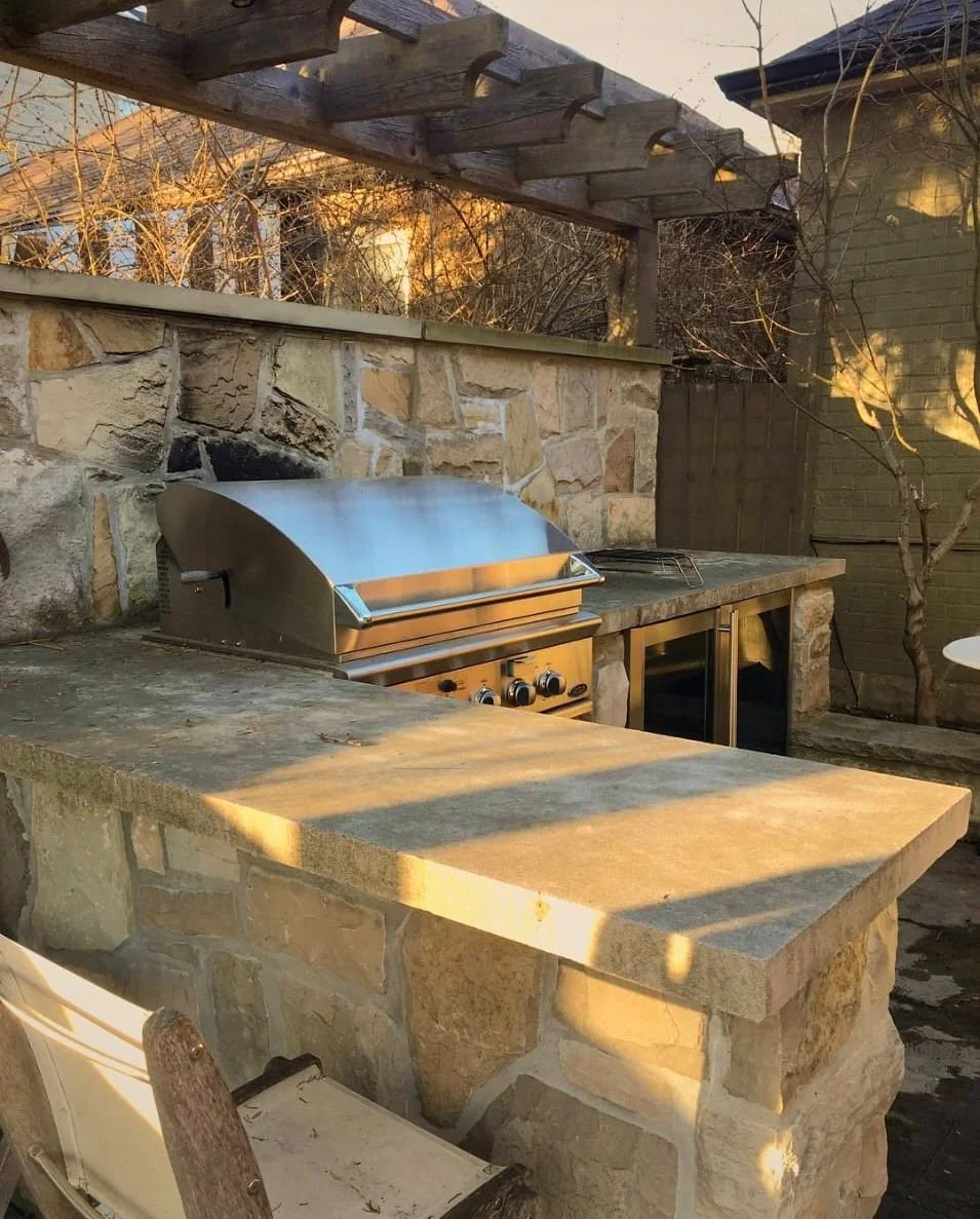 Outdoor kitchen with a stainless steel grill, stone countertop, and stone wall, surrounded by a wooden pergola and bare tree branches.