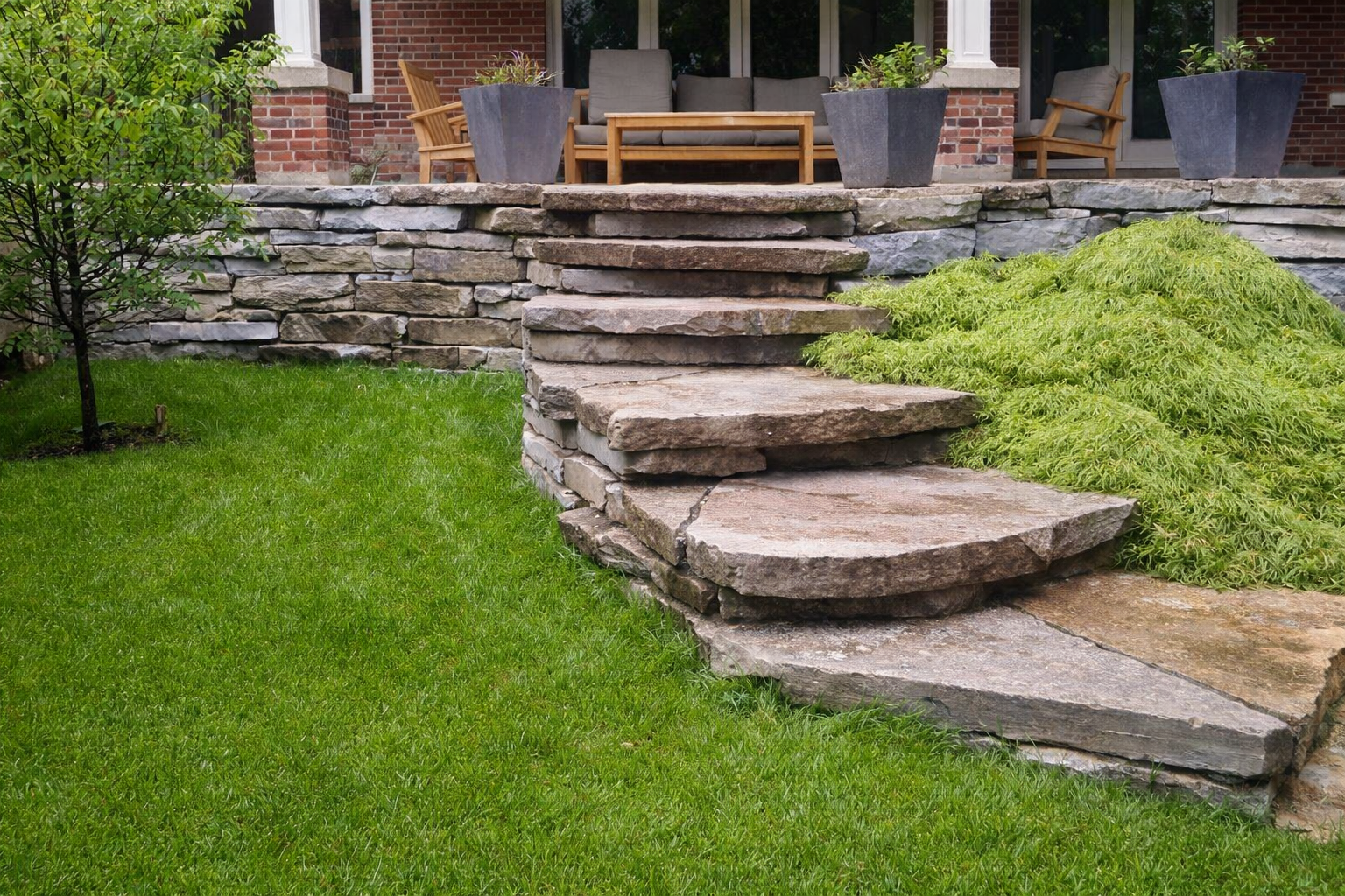 Stone steps leading up to a brick patio with outdoor furniture and potted plants, surrounded by a lush green lawn.