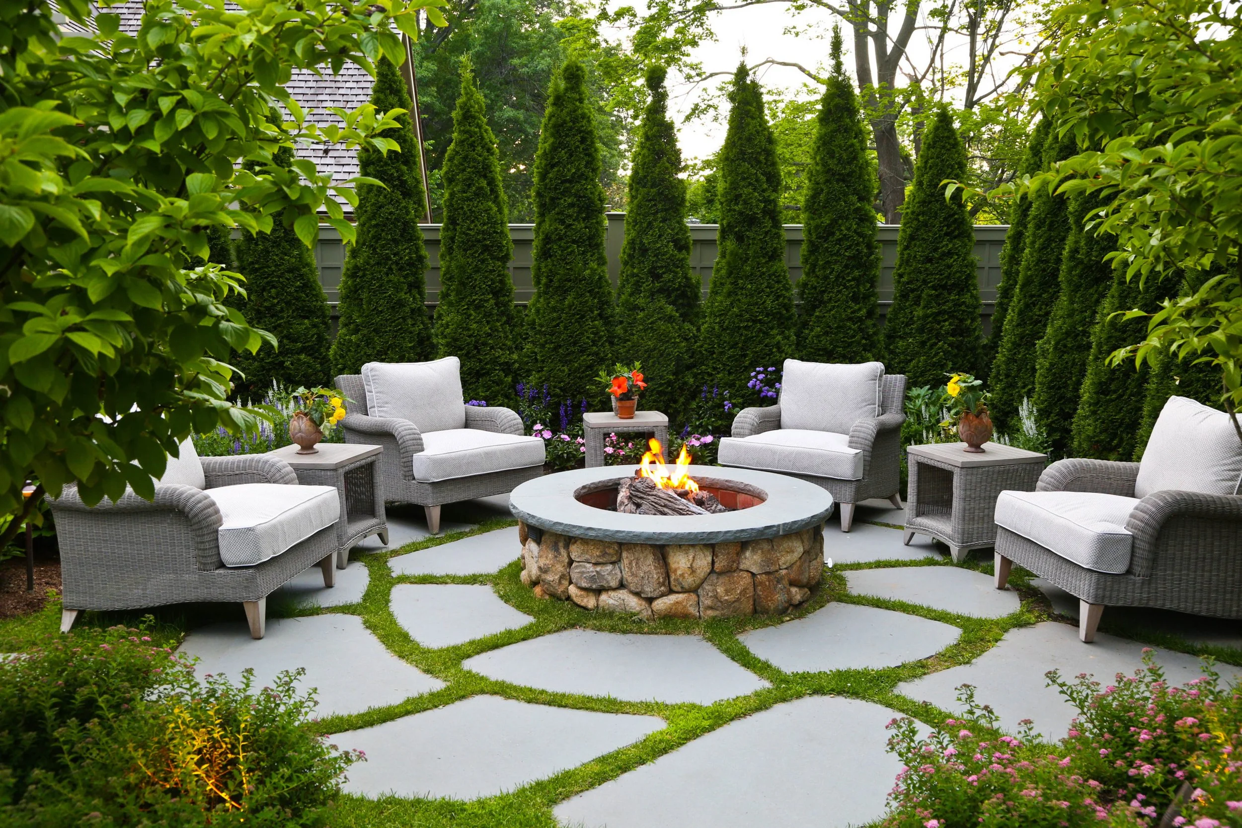 Outdoor seating area with six gray wicker chairs around a stone fire pit, surrounded by a lush green garden with tall trees and flowering plants.