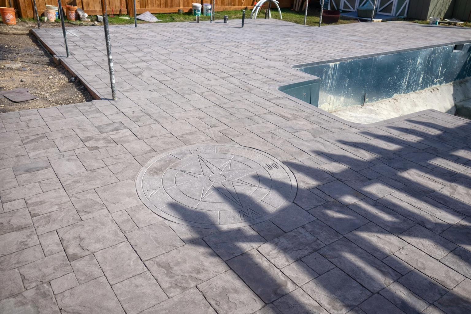 A newly paved backyard with brick-patterned concrete, a compass rose design on the ground, a pool under construction, and a wooden fence in the background.