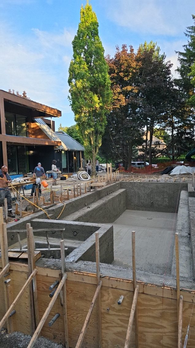 Construction site with a swimming pool foundation being poured, surrounded by wooden framing and workers, with trees and a building in the background.