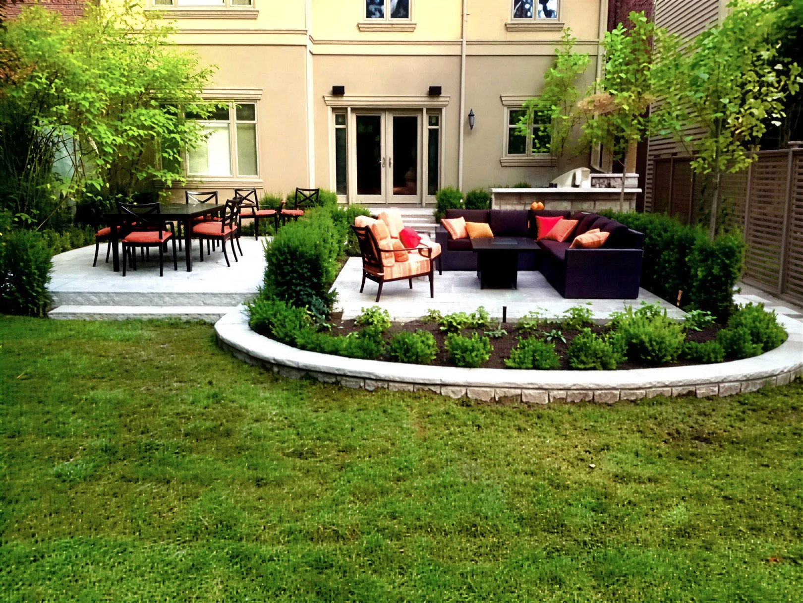 A backyard patio with outdoor furniture, including a circular black sofa with yellow and orange cushions, a single armchair with yellow and red cushions, and a rectangular table. To the left, there is a dining area with a round table and six chairs w