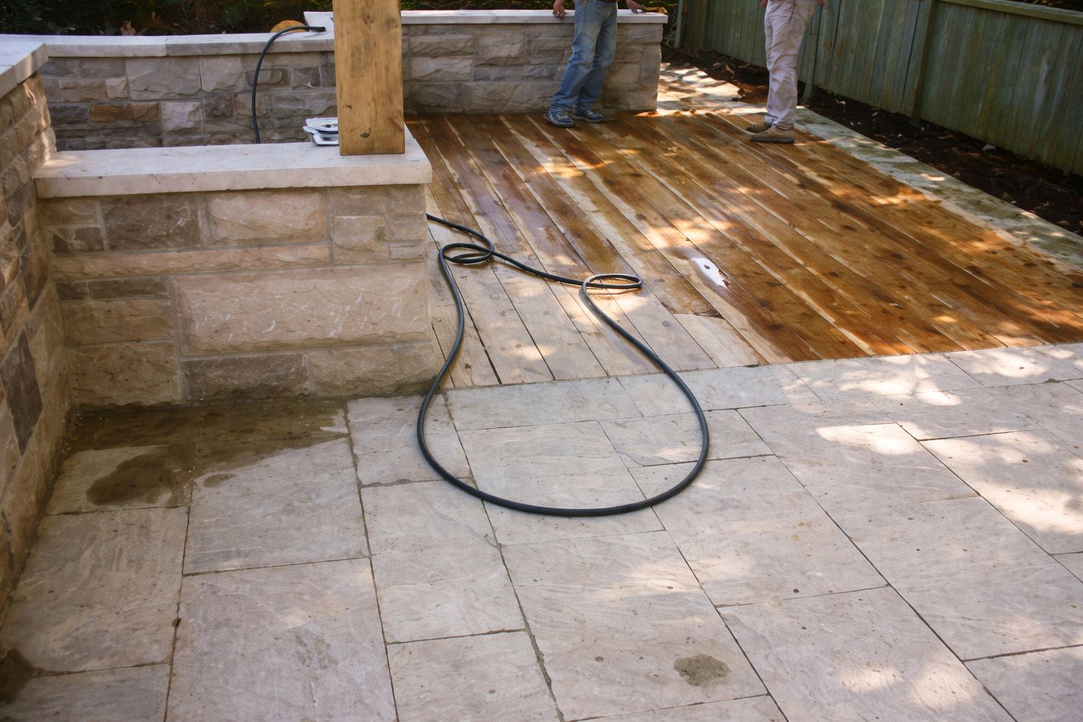 Outdoor patio area showing a partially installed wooden deck, stone paving, and a garden hose on the ground, with two people working in the background.