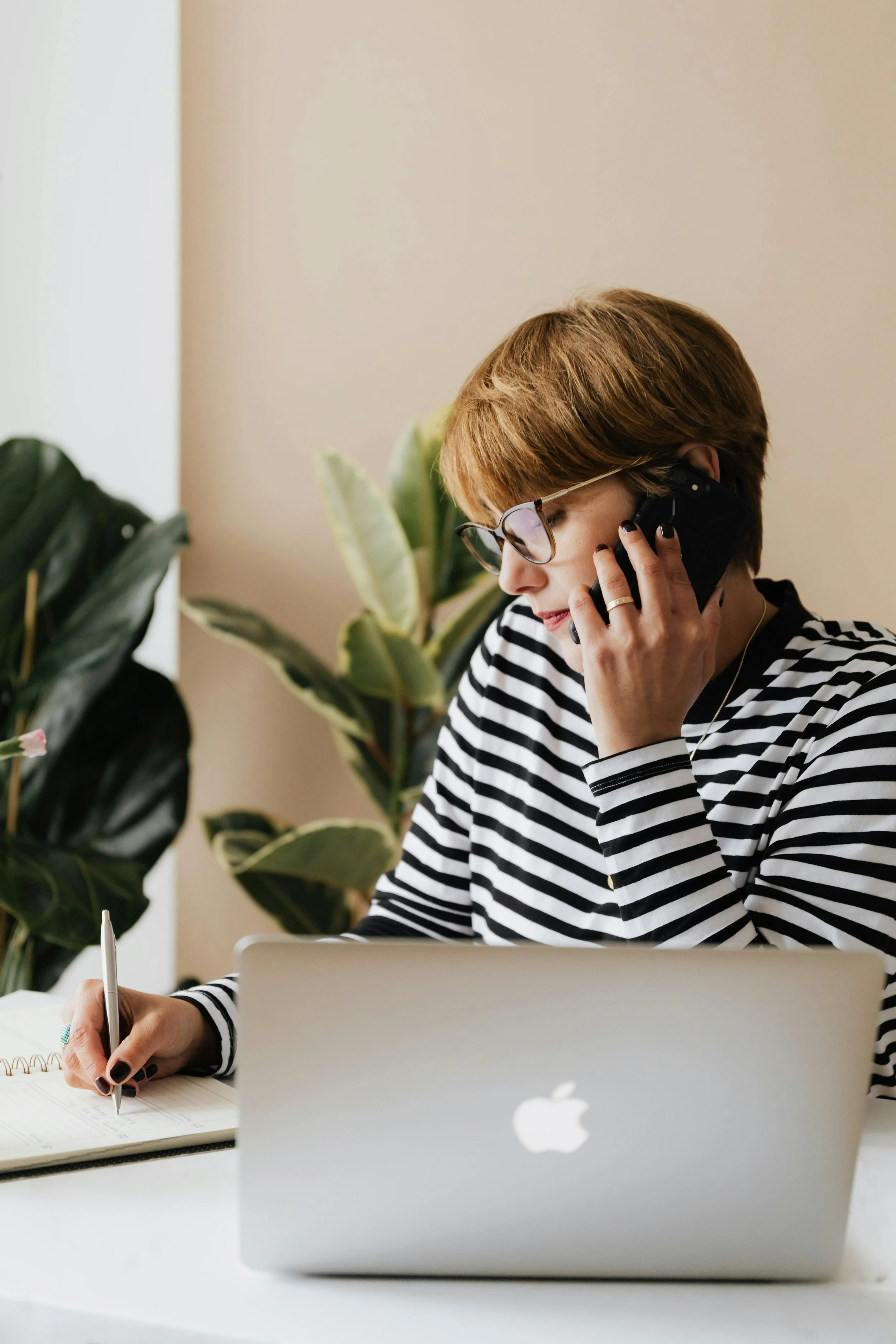 A woman with short hair and glasses, wearing a black and white striped shirt, talking on a phone while writing in a notebook with a pen. She is sitting at a desk with a silver MacBook in front of her, and there are green plants in the background.