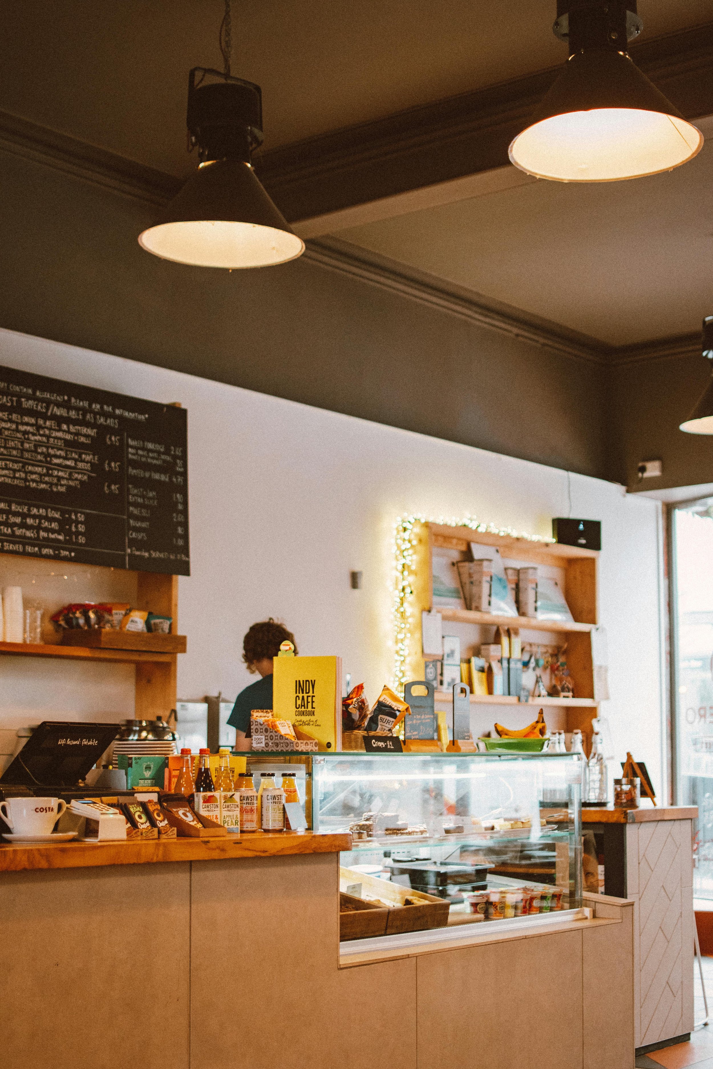 Interior of a cozy coffee shop with a counter displaying snacks and a glass display case for baked goods. There are shelves with books and decorative lights on the wall behind the counter. Overhead pendant lights illuminate the space.