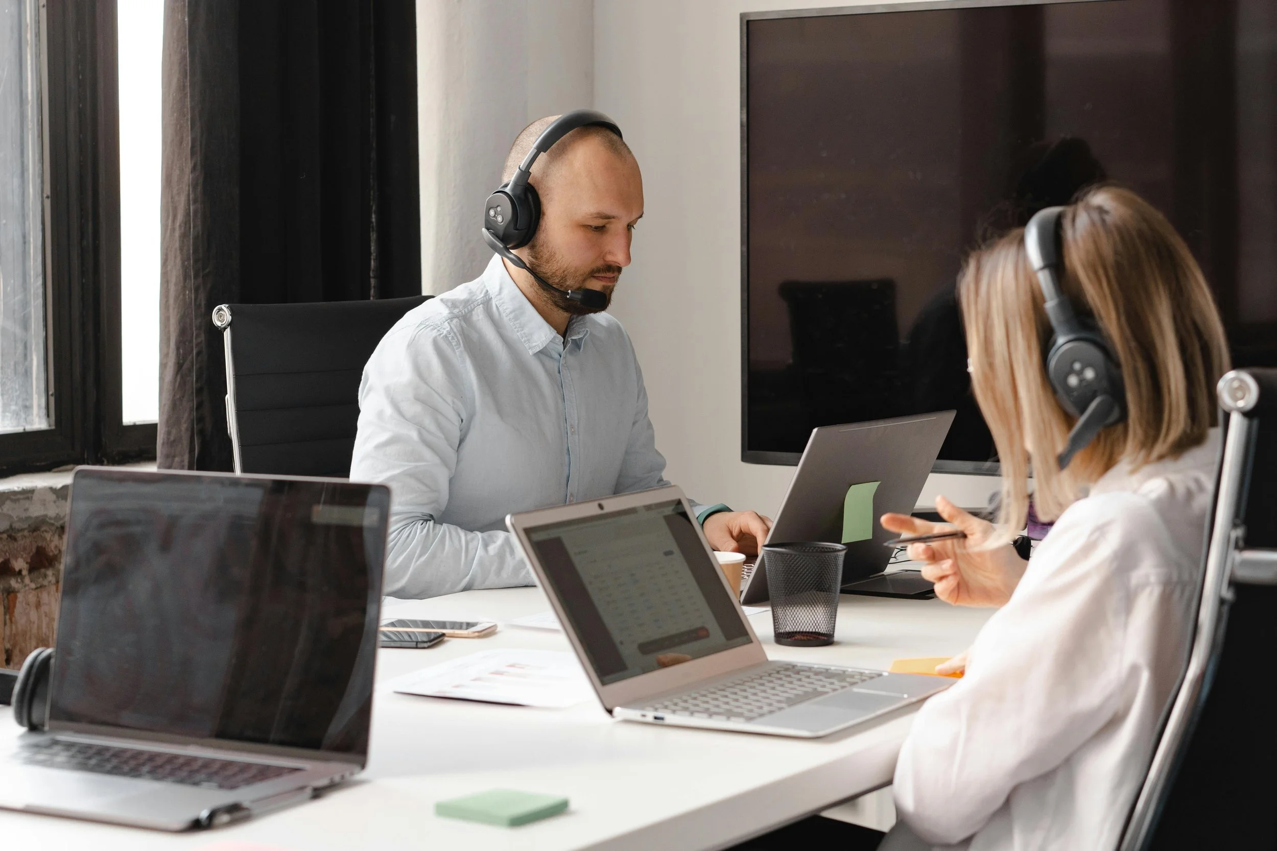 Two people sitting at a white table in an office, wearing headsets, working on laptops, with a large monitor and various electronic devices on the table.
