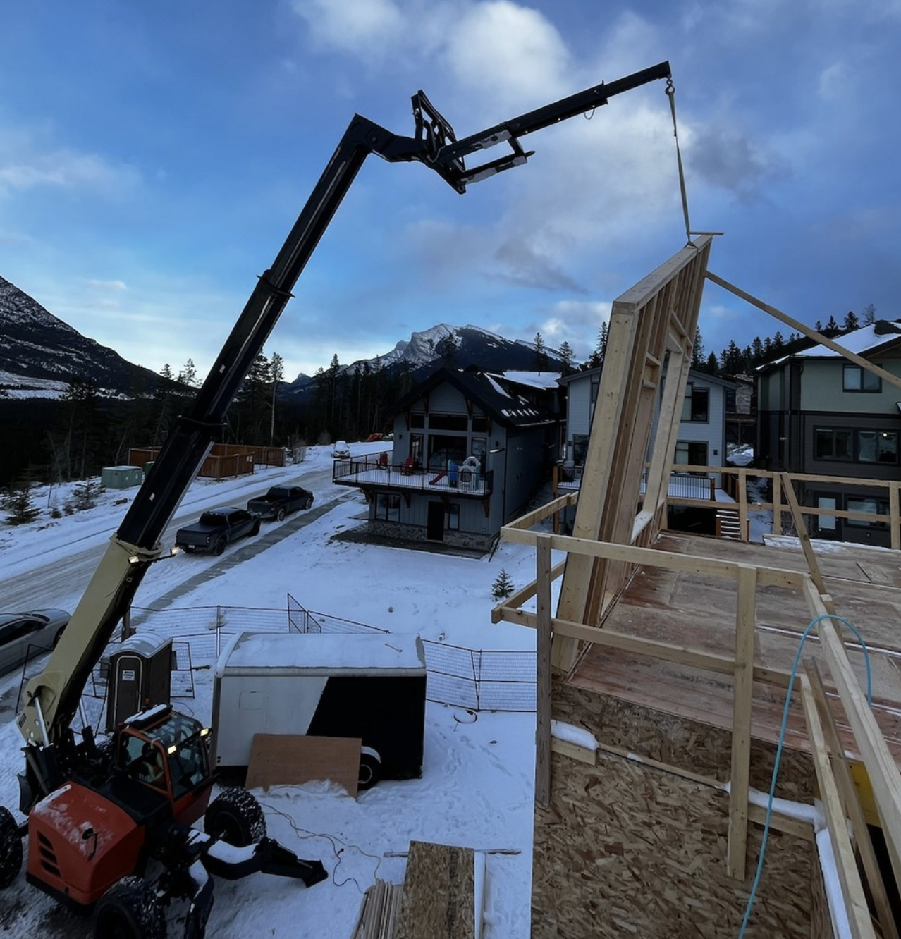 Construction site in a snowy residential area with a crane lifting a wooden wall section, surrounded by houses and mountain scenery.