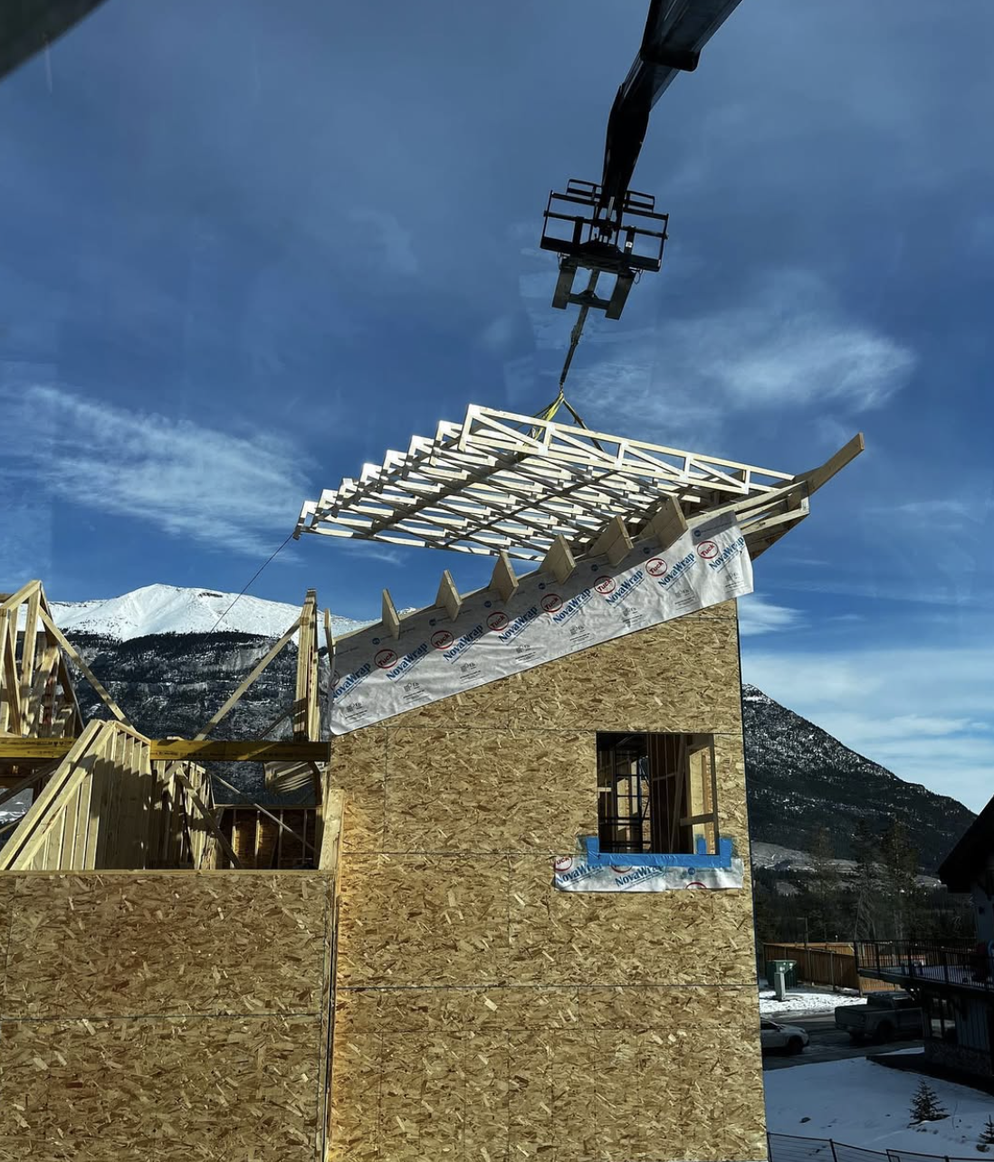 Construction site of a house with wooden framing and partially installed roof, with a crane lifting roofing materials against a mountainous background and blue sky.