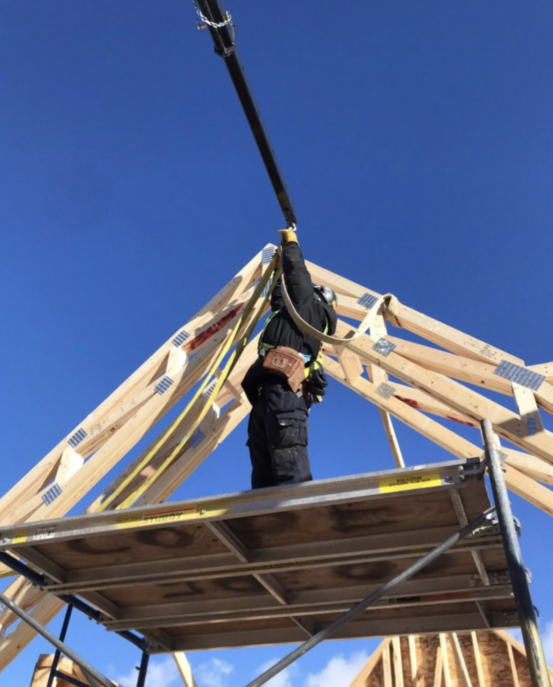 A construction worker wearing safety gear, including a helmet and harness, working on a wooden framework of a building under a clear blue sky.