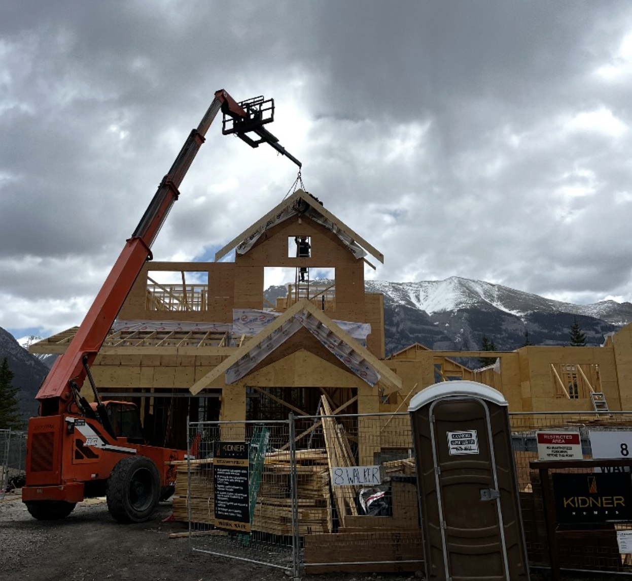 Construction of a wooden house with a crane lifting roofing materials, mountains in the background, and cloudy sky overhead.