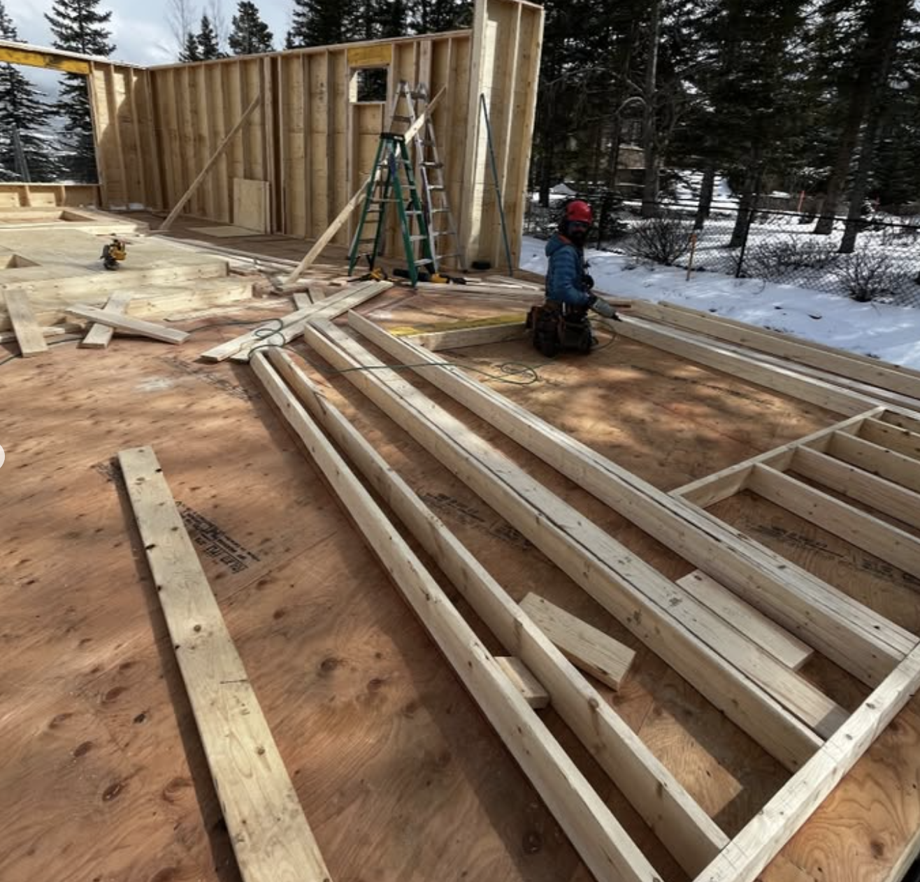 Construction site with wooden framing, a worker in a red helmet kneeling among scattered lumber, surrounded by trees and snow.