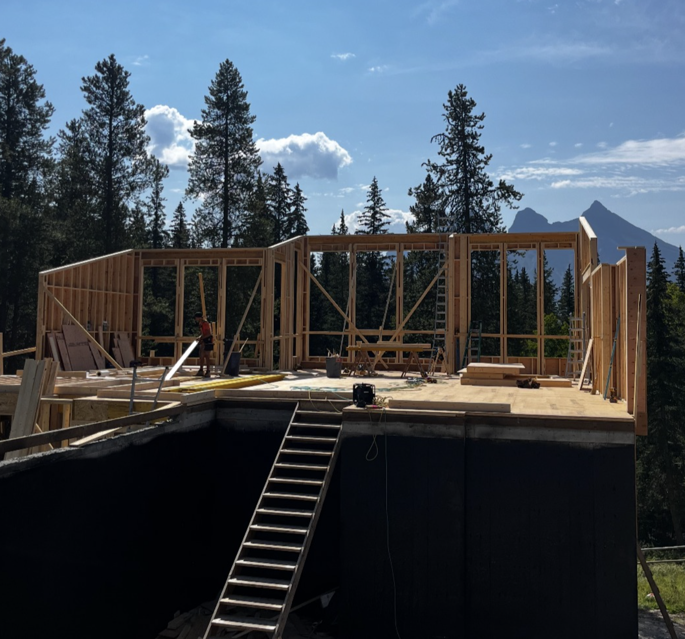 Wooden framing of a house under construction on a raised platform surrounded by trees and mountains in the background.