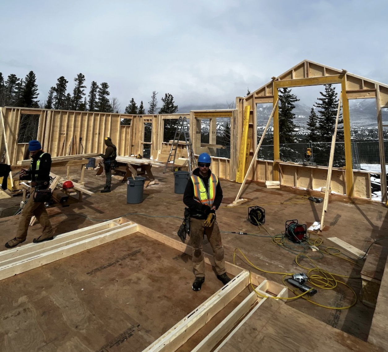Construction workers building a wooden framework for a house in a snowy, mountainous landscape with trees in the background.
