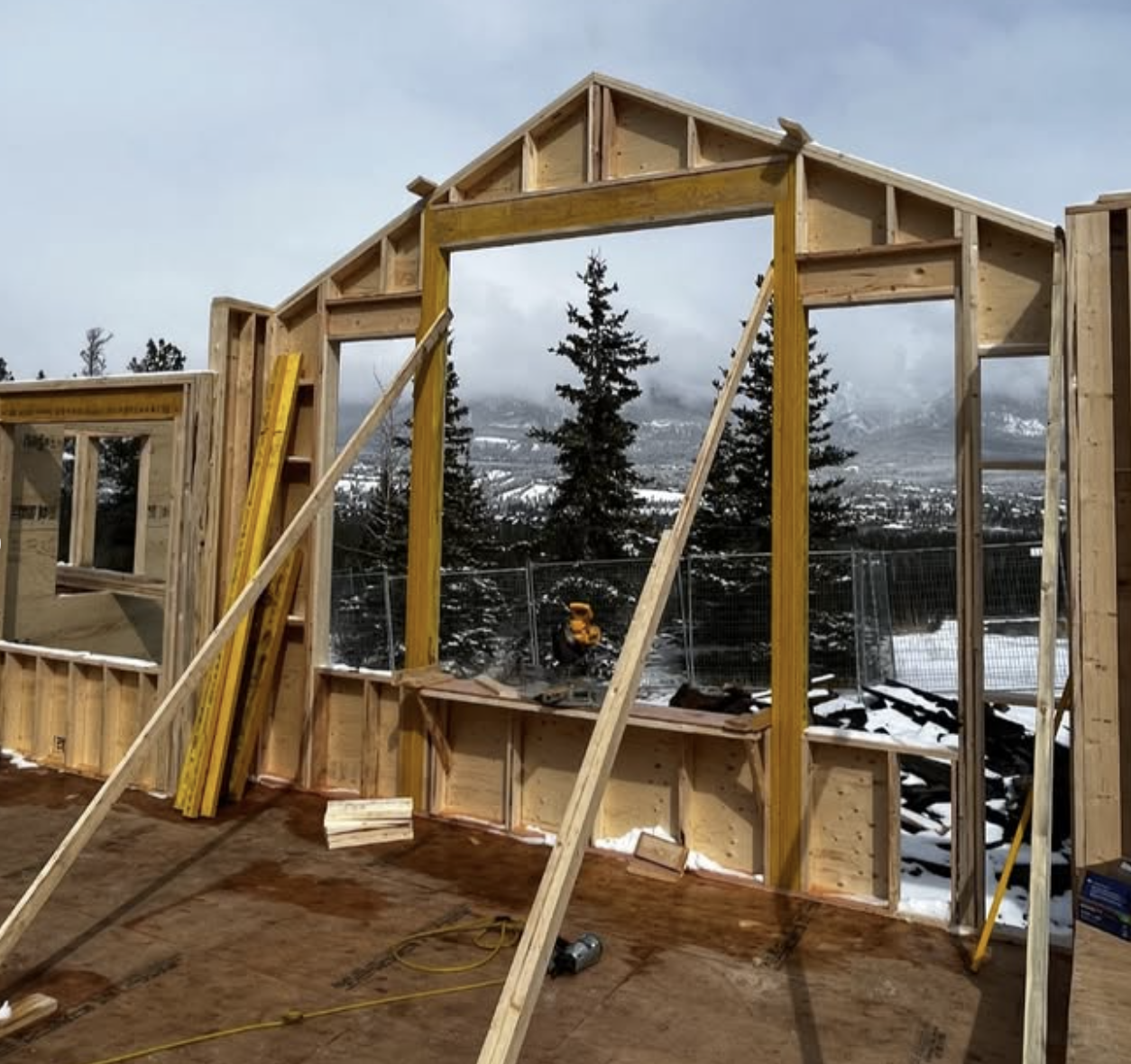 Construction site with wooden framing of a building under construction, snow on the ground, trees, and mountains in the background.