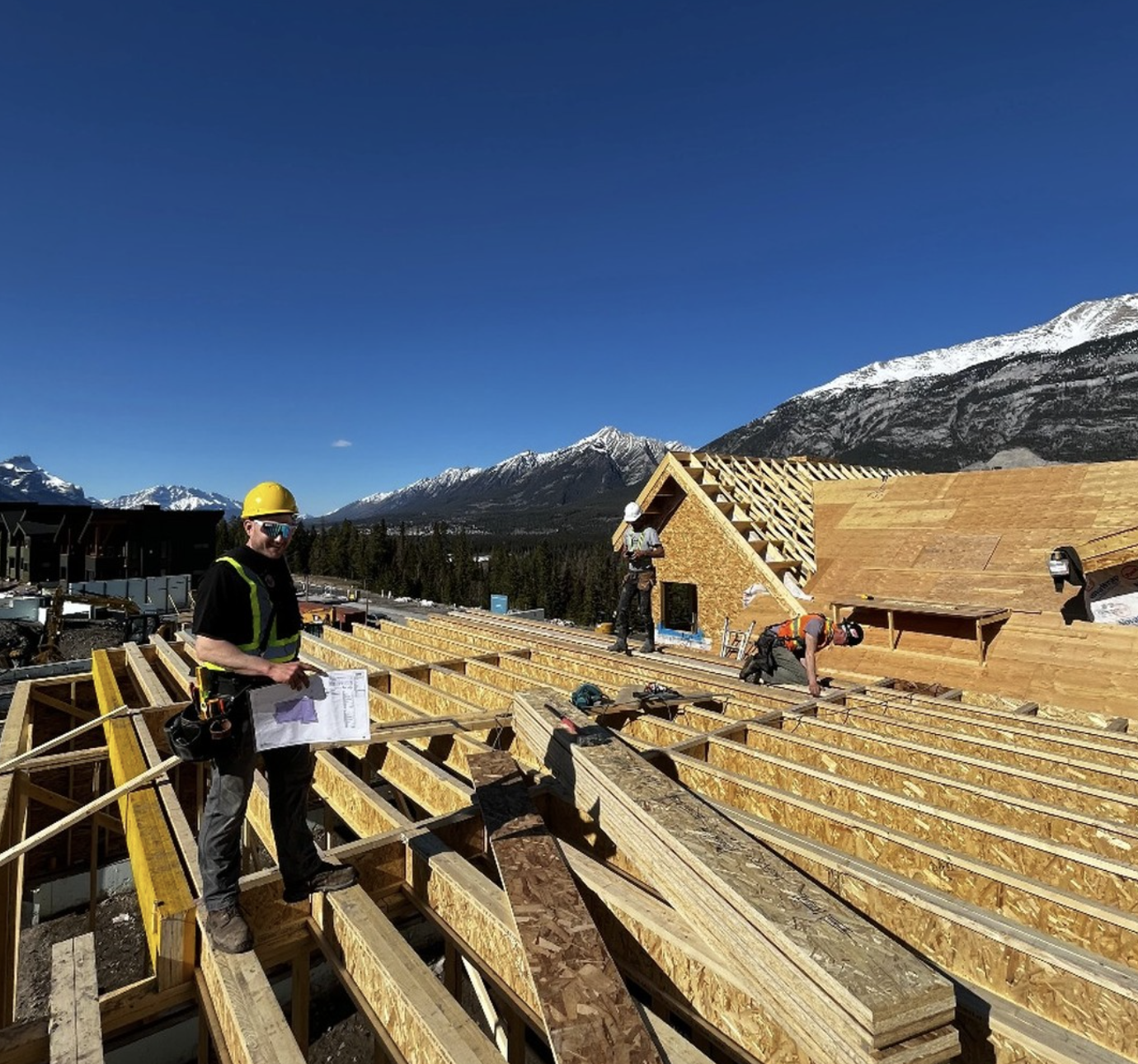 Construction workers building a wooden house in a mountainous area with snow-capped peaks under a clear blue sky.