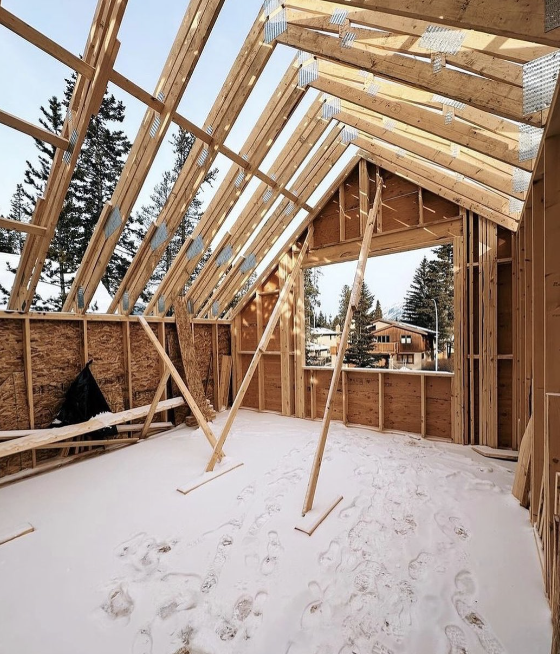 The interior of a house under construction with exposed wooden framing, snow on the floor, and footprints visible. The view shows the roof structure and large openings for windows, with neighboring houses and tall pine trees outside.