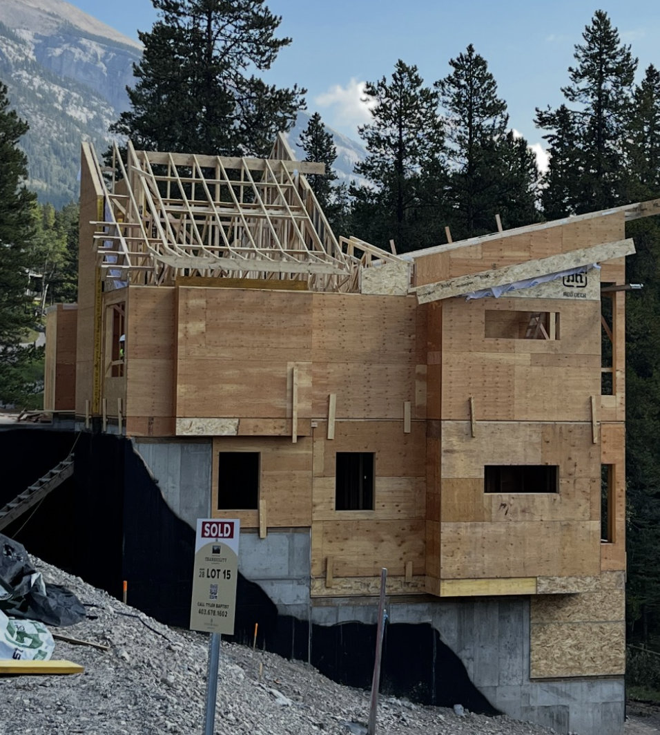 A wooden house under construction, situated on a concrete foundation, with trees and mountains in the background. Building materials and a 'sold' sign are visible in the foreground.