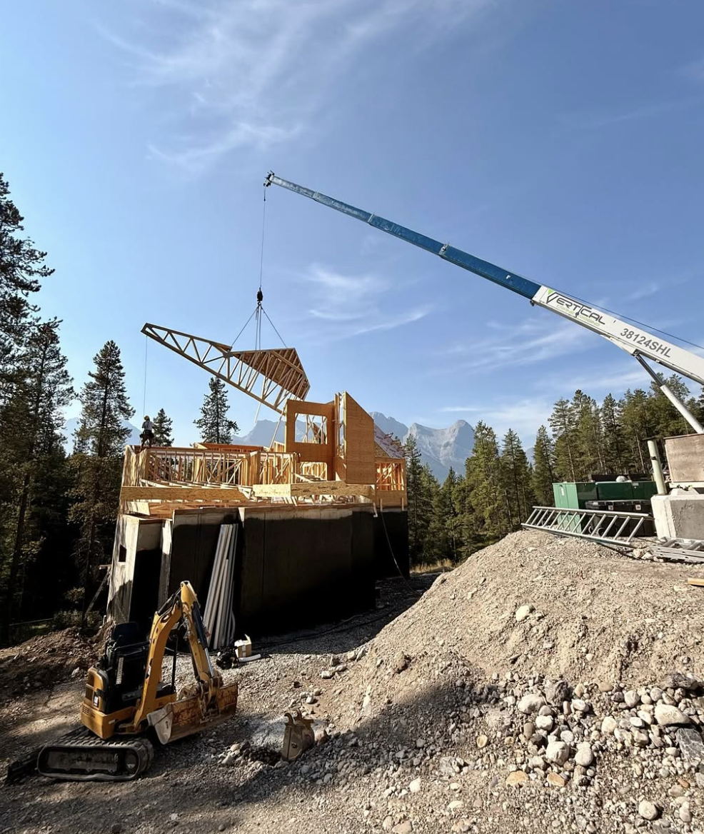 Construction site with a crane lifting a wooden framework structure, surrounded by trees and mountain scenery.