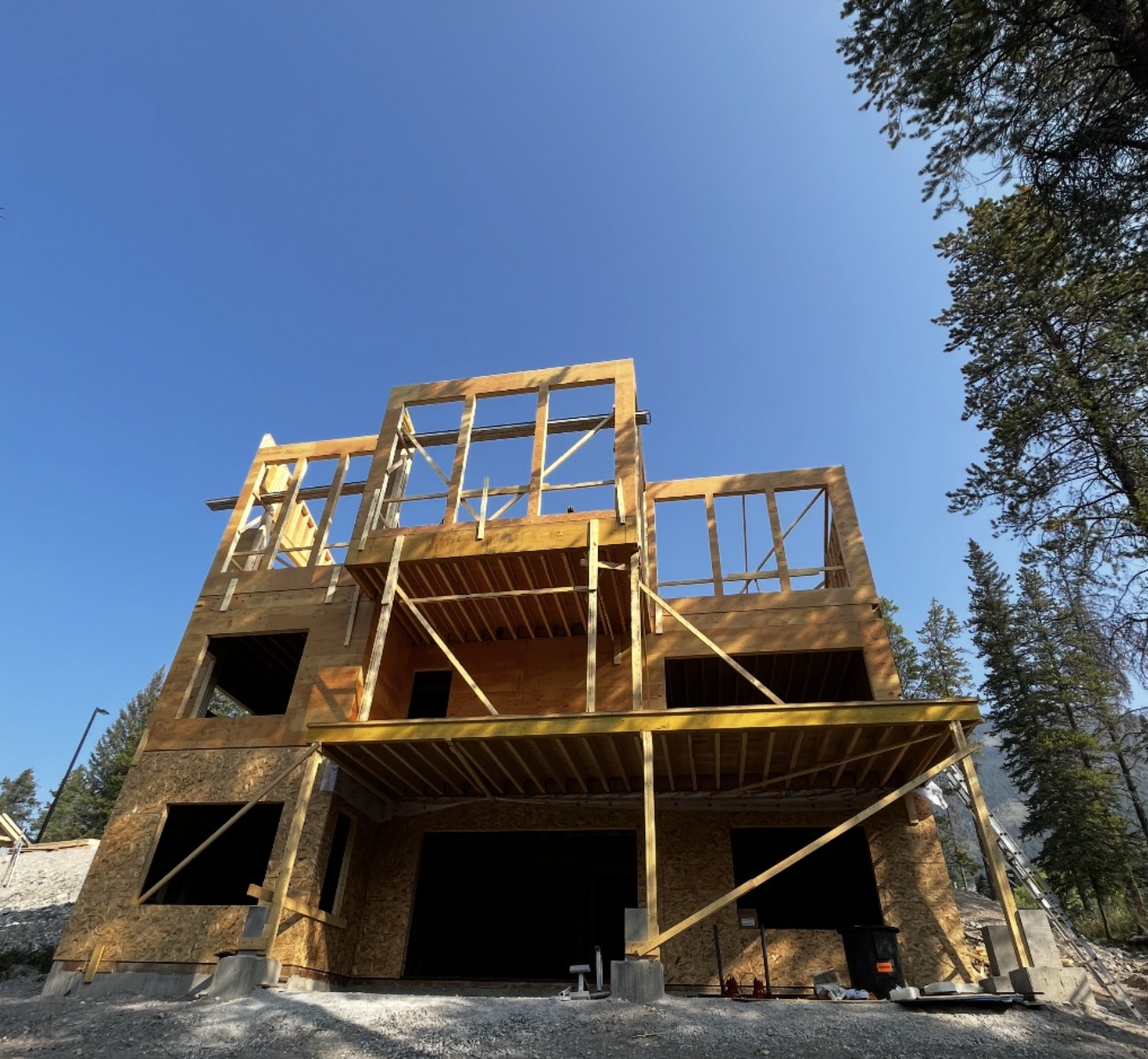 A multi-story house under construction with a wooden frame, situated on a gravel lot surrounded by trees, against a clear blue sky.