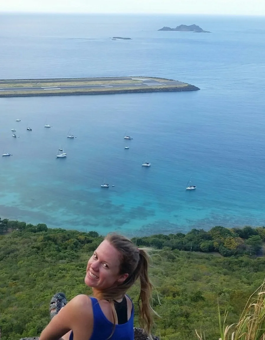 A woman smiling and sitting on a hillside overlooking a coastal area with boats anchored in the turquoise water and an island in the distance.