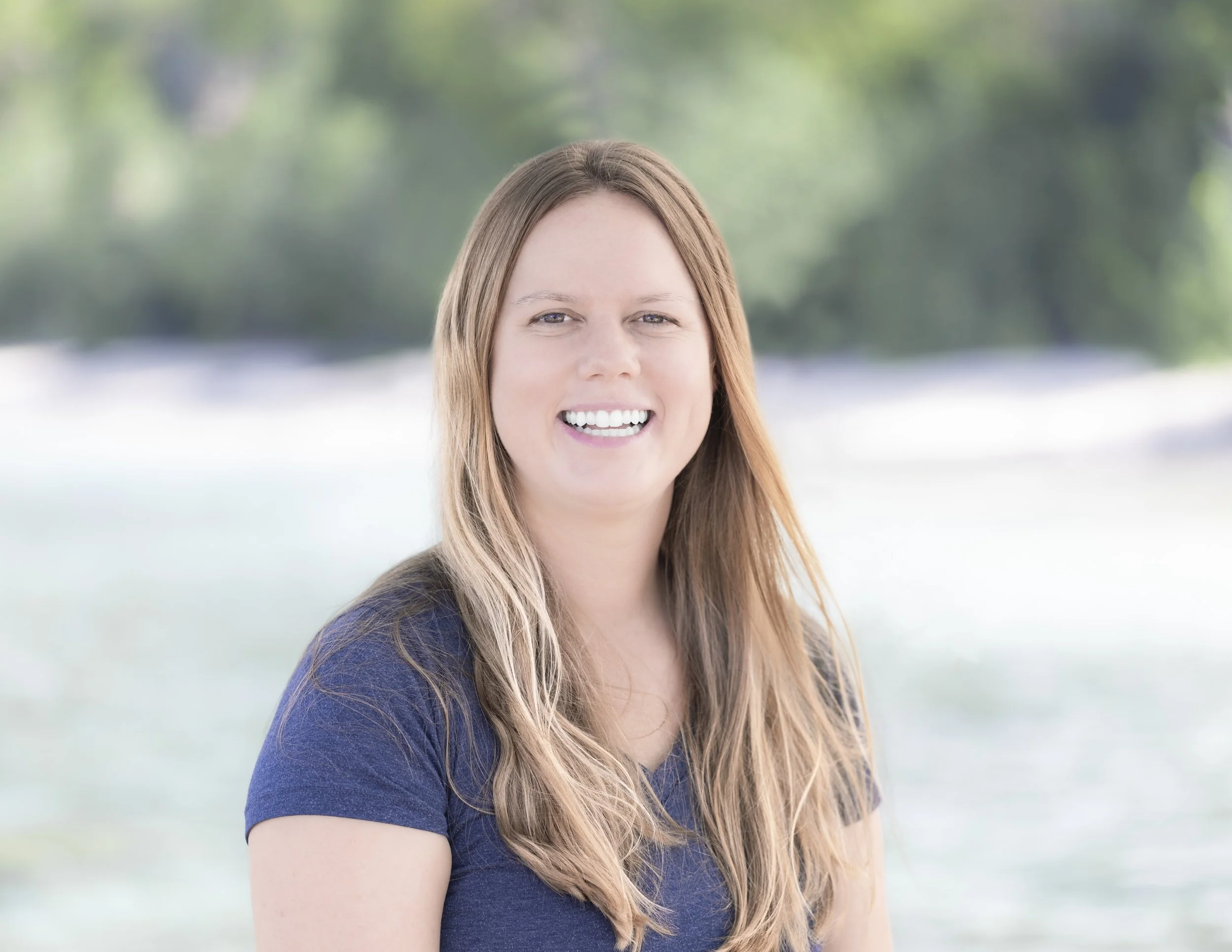 A woman with long, light brown hair smiling outdoors with a blurred natural background.