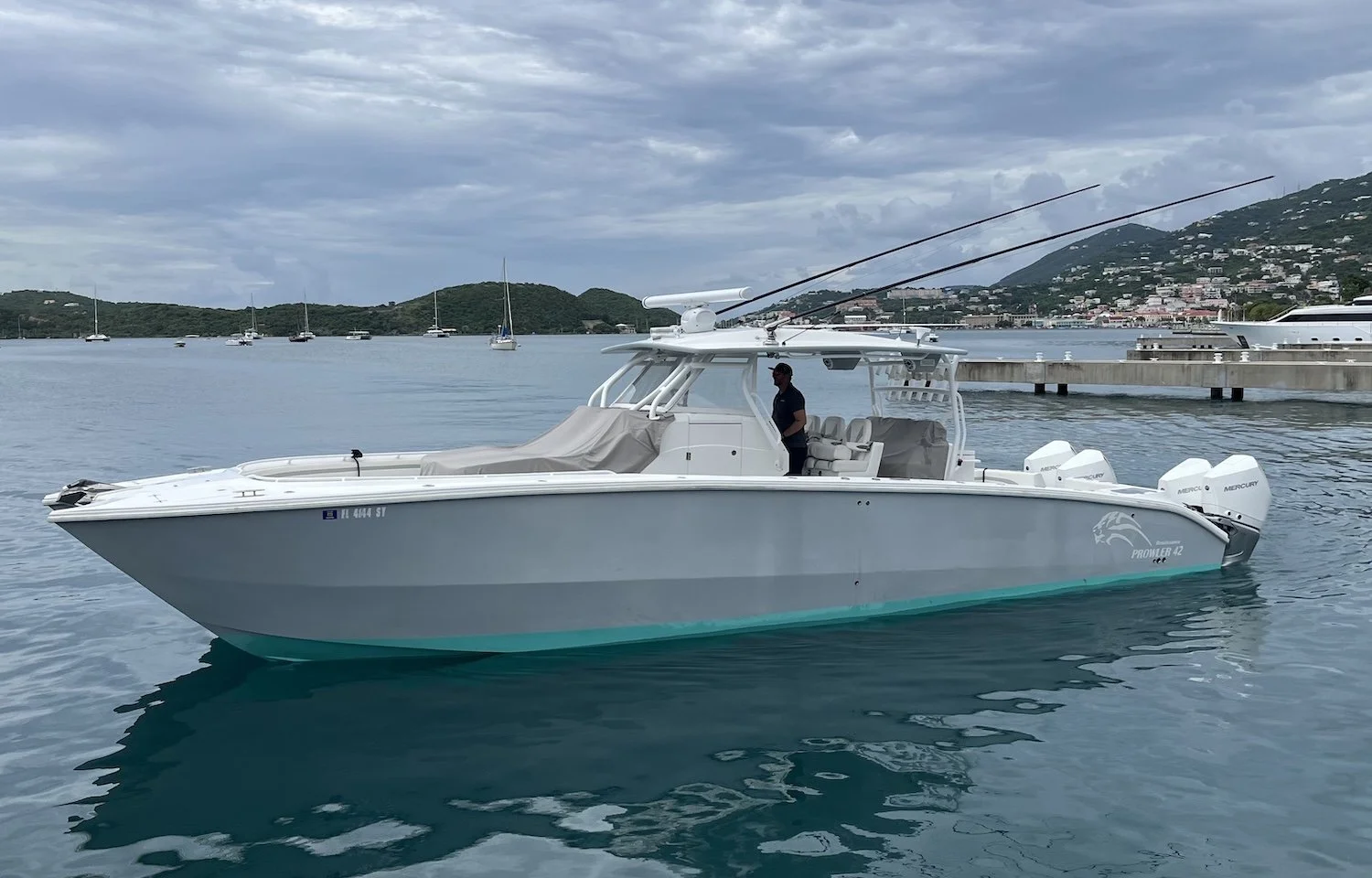 A white fishing boat with seafoam-colored bottom, floating on calm water near a harbor with other boats and a hilly landscape in the background.