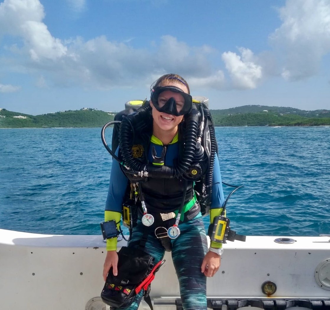A woman in scuba gear standing on a boat, smiling, with a blue ocean, green hills, and partly cloudy sky in the background.