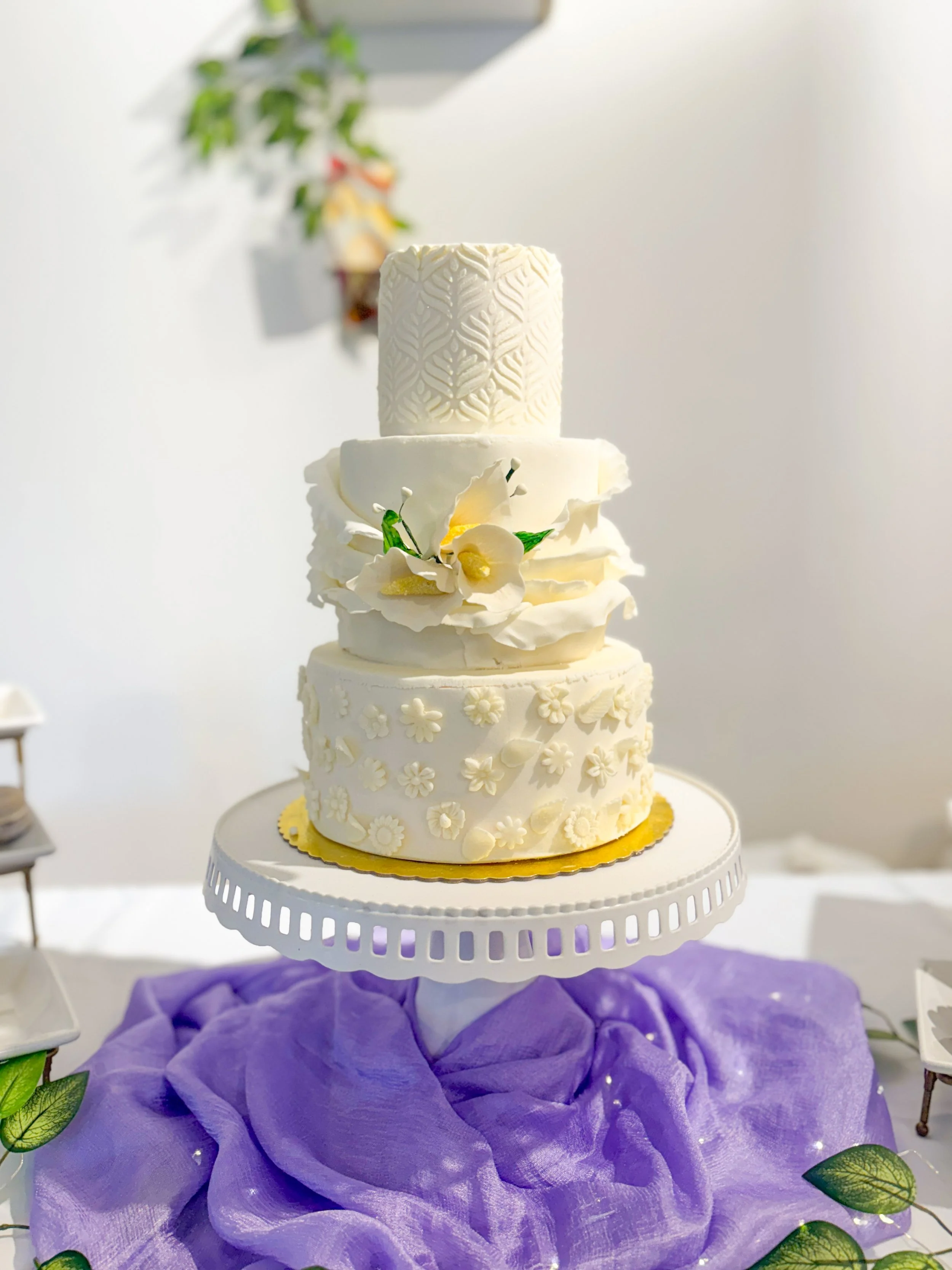 A multi-tiered white wedding cake decorated with intricate floral and leaf patterns, topped with a cylindrical design, and surrounded by purple fabric and greenery.