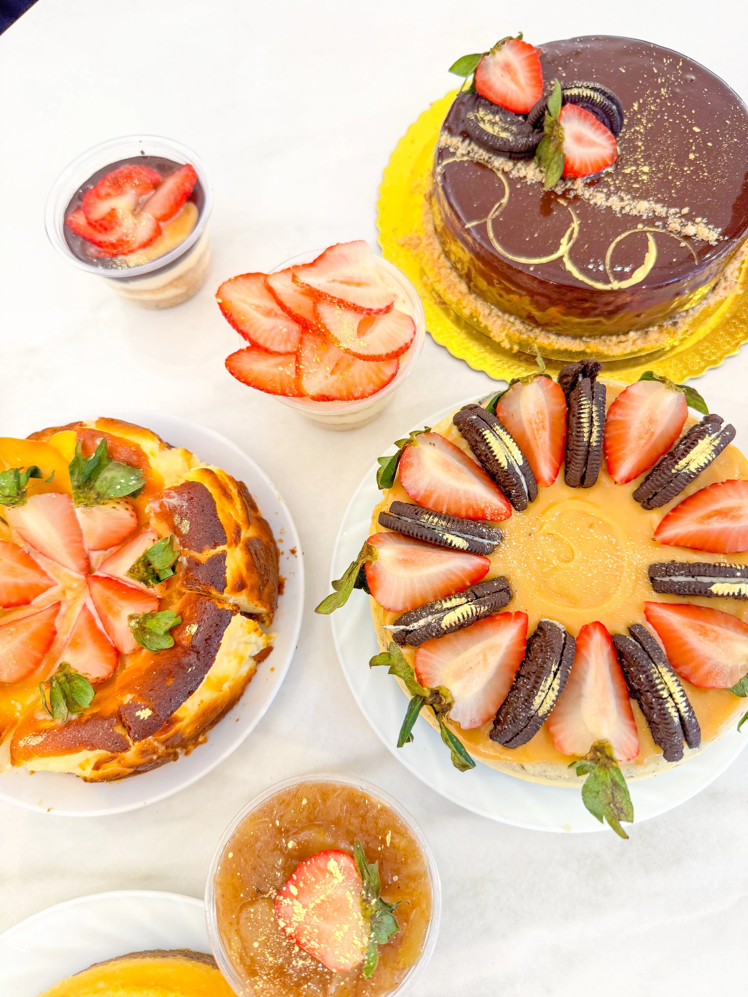 Assorted desserts, including a large chocolate-glazed cake garnished with strawberries and Oreos, and small pudding cups with strawberries, on a white surface.