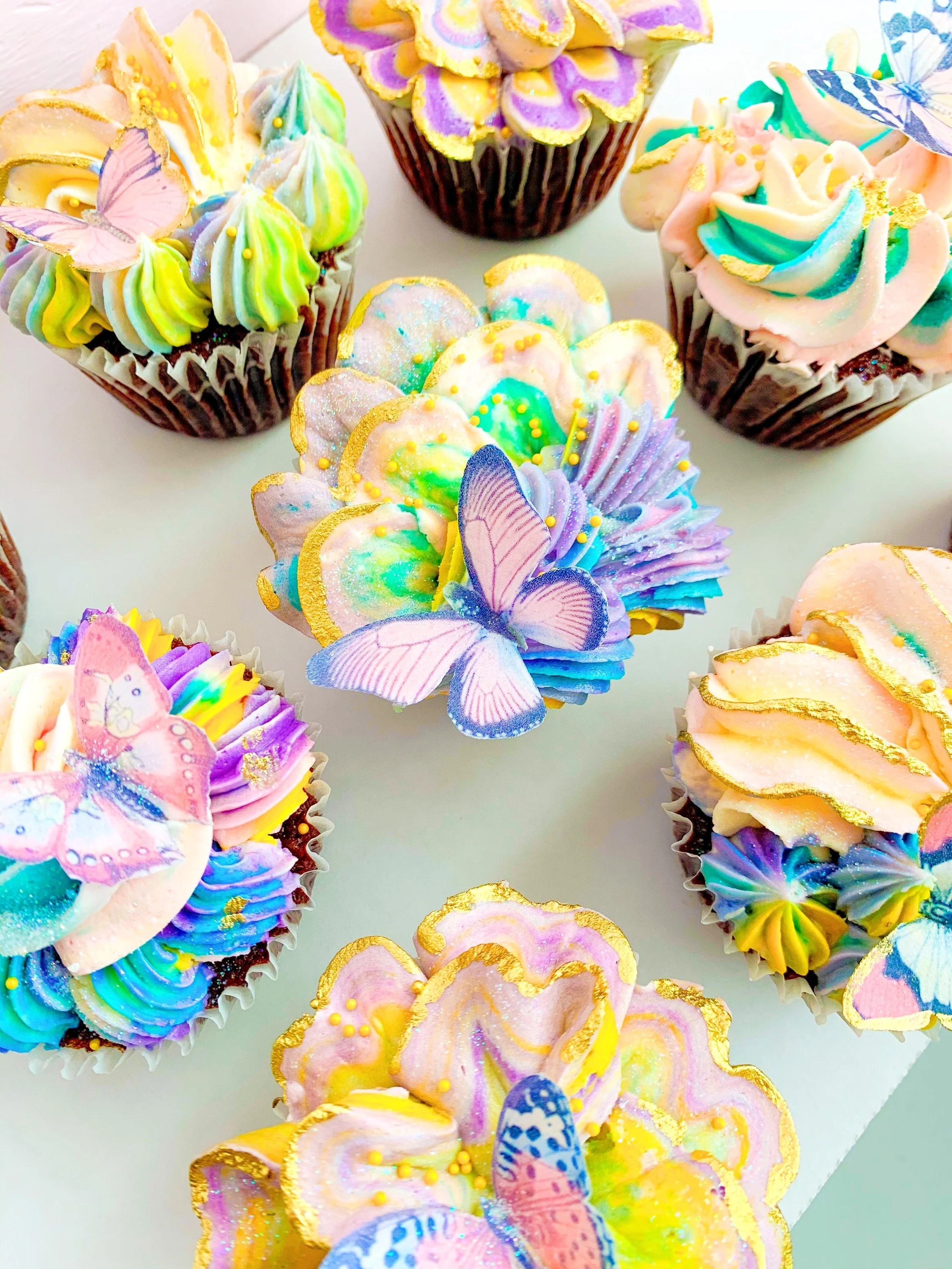 Colorful cupcakes decorated with pastel swirls, strawberries, butterflies, and sprinkles, topped with gold accents.