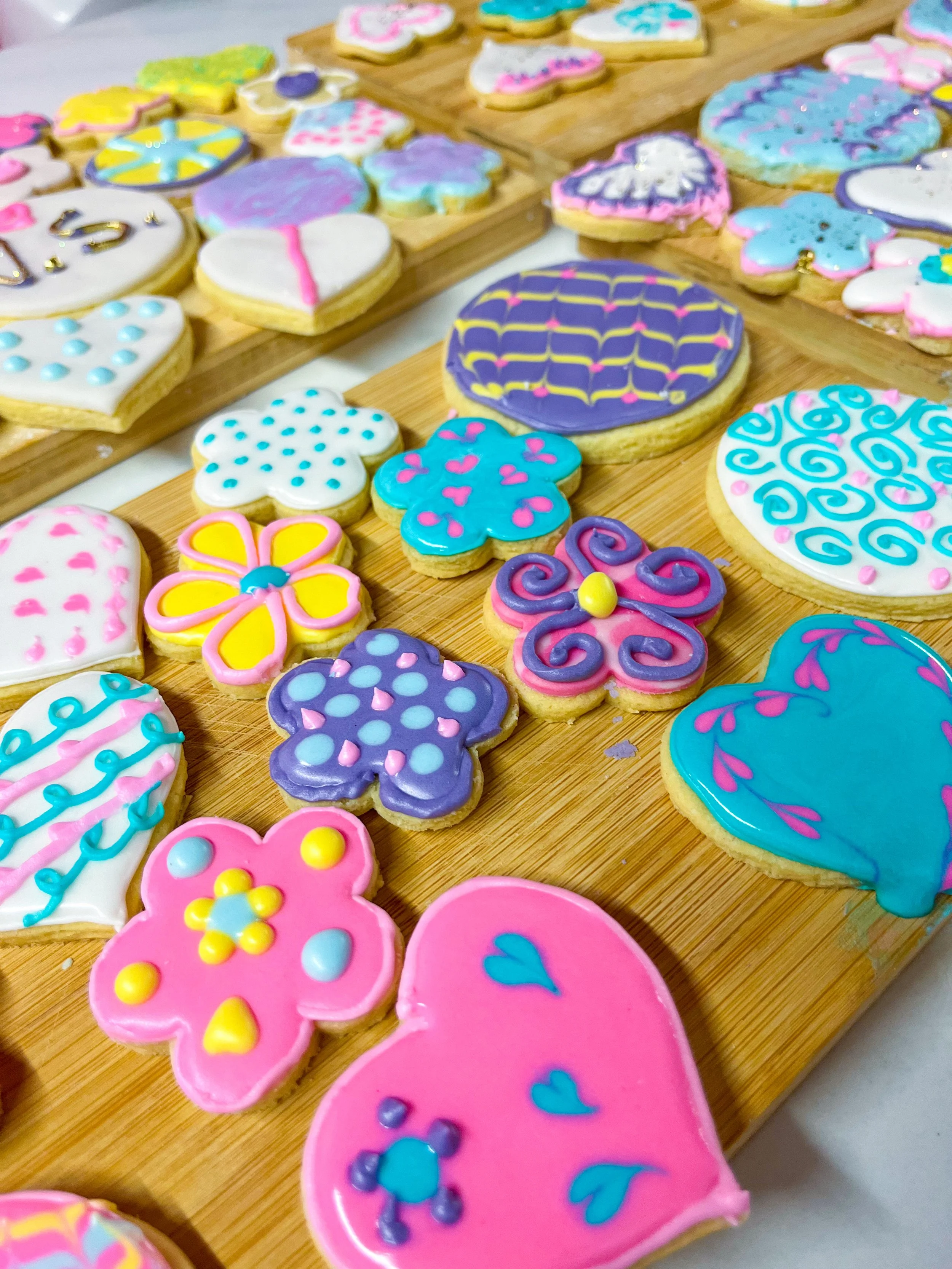 Colorful decorated sugar cookies in various shapes, including hearts, butterflies, and flowers, with pink, purple, blue, yellow, and white icing and decorative accents, displayed on wooden boards.