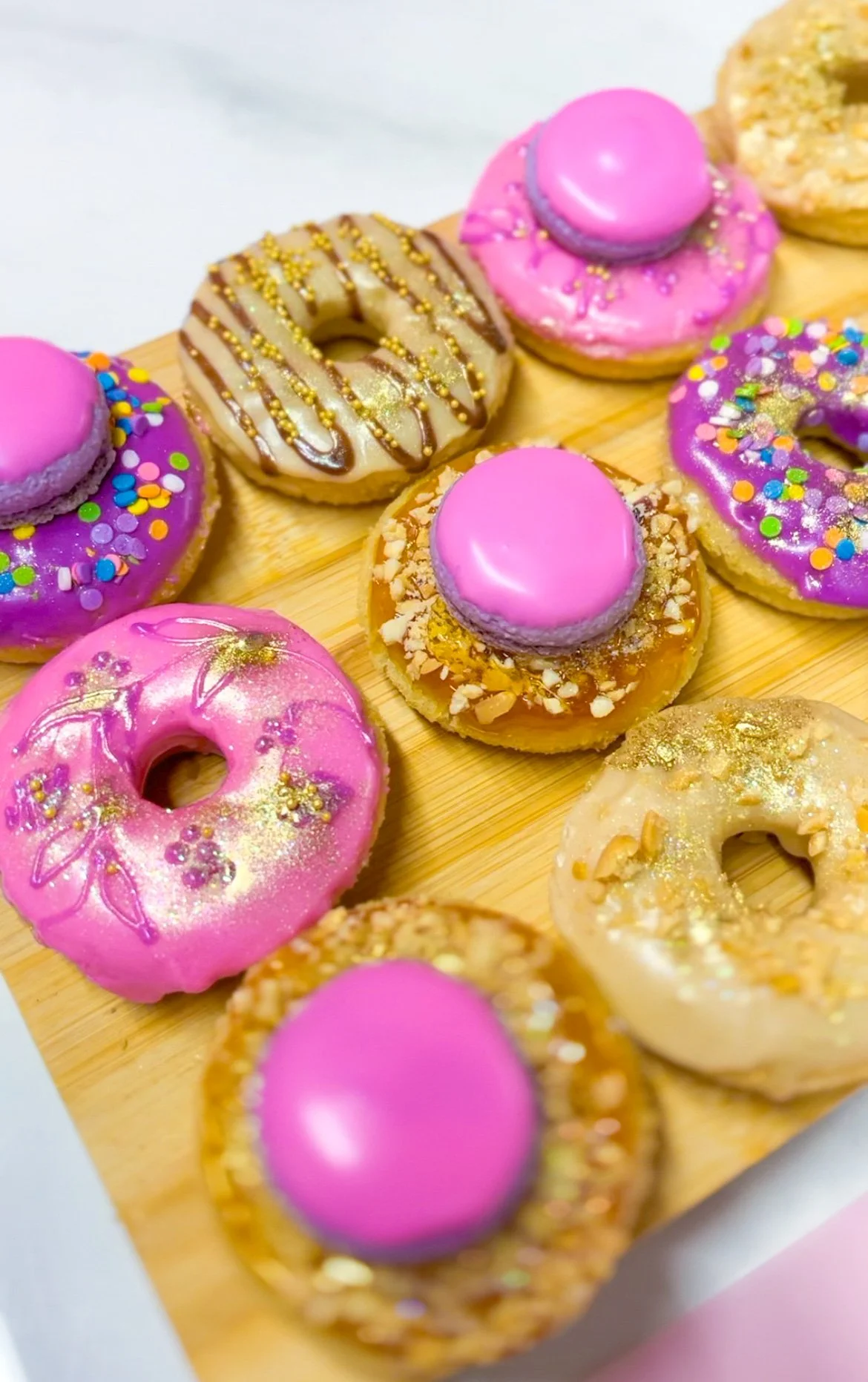 Colorful assorted donuts with pink, purple, white frosting, sprinkles, and toppings on a wooden tray.