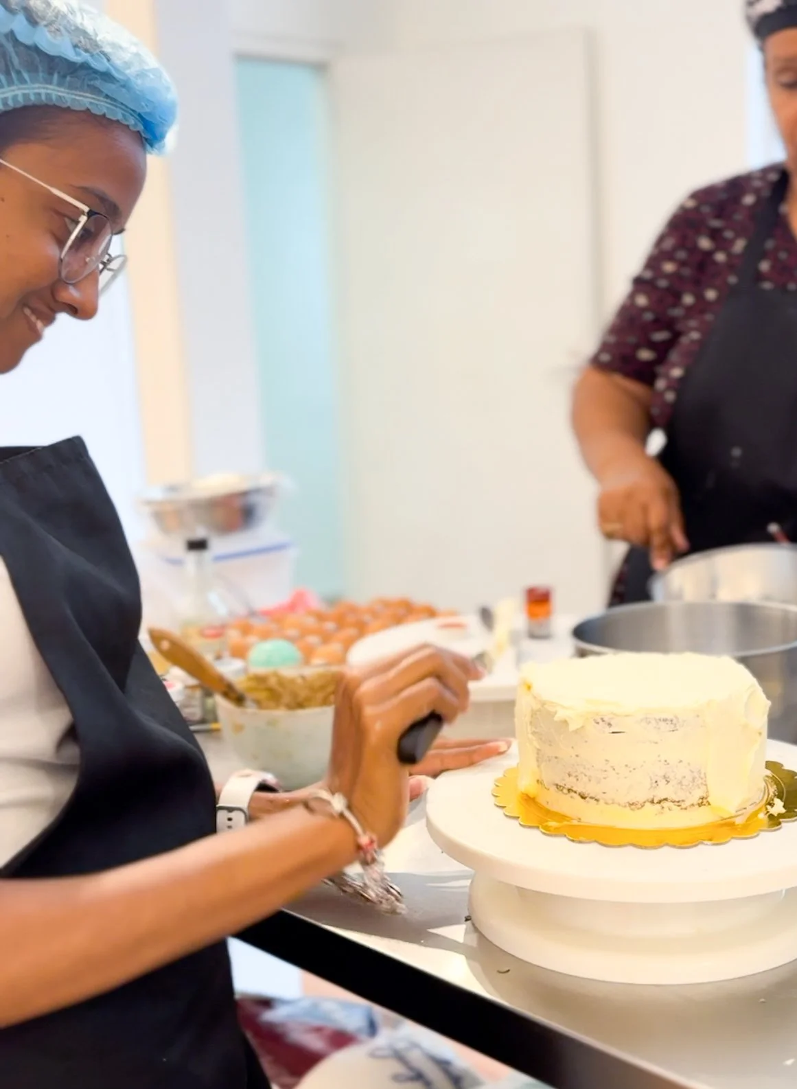 People decorating a cake in a kitchen or bakery, with baking ingredients and supplies on the counter.