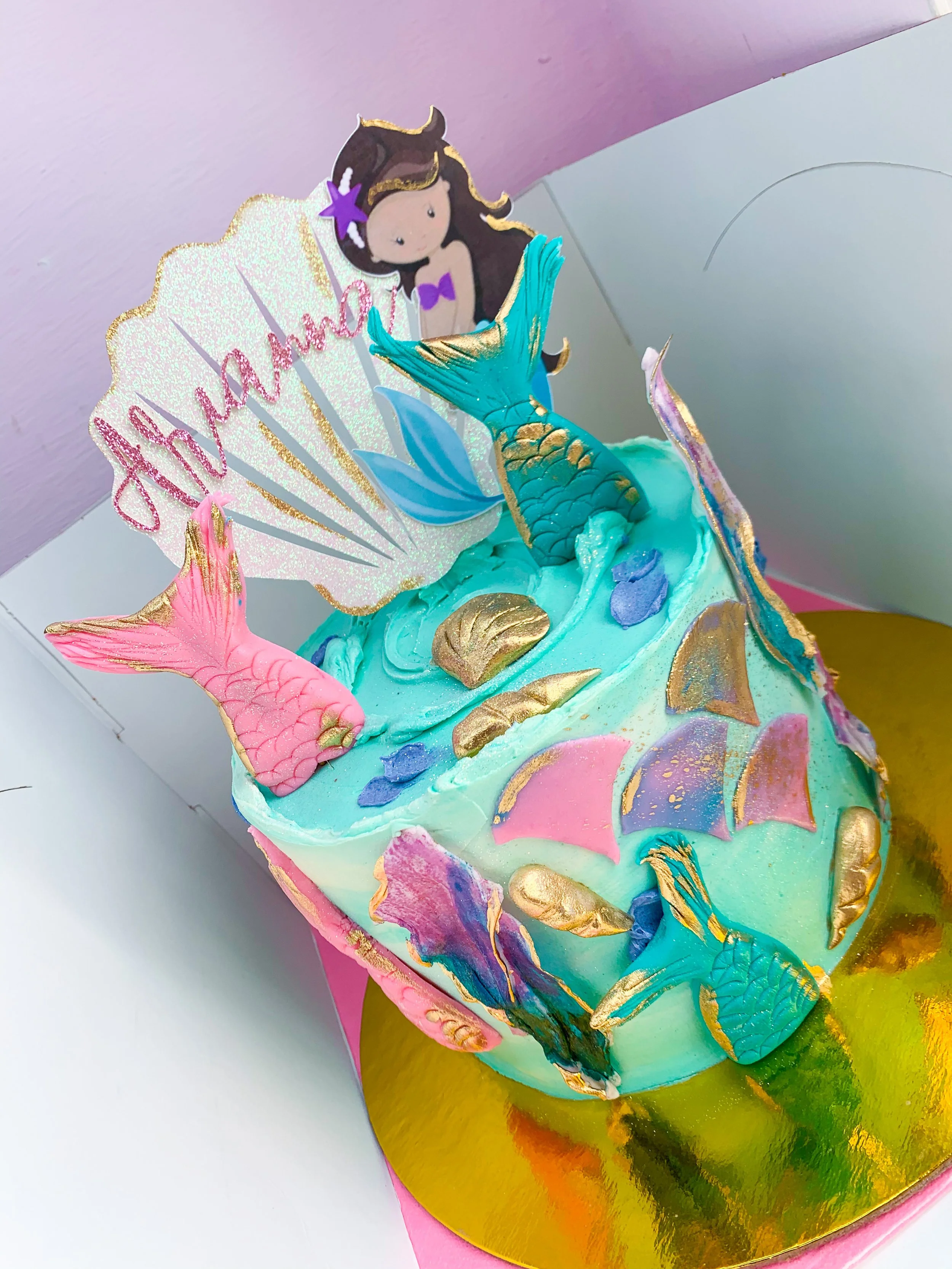 A birthday cake decorated with a mermaid theme, featuring a mermaid topper, colorful seashell and starfish decorations, and a 'Happy Birthday' sign. The cake has pastel-colored icing and metallic gold accents, placed on a reflective gold base.